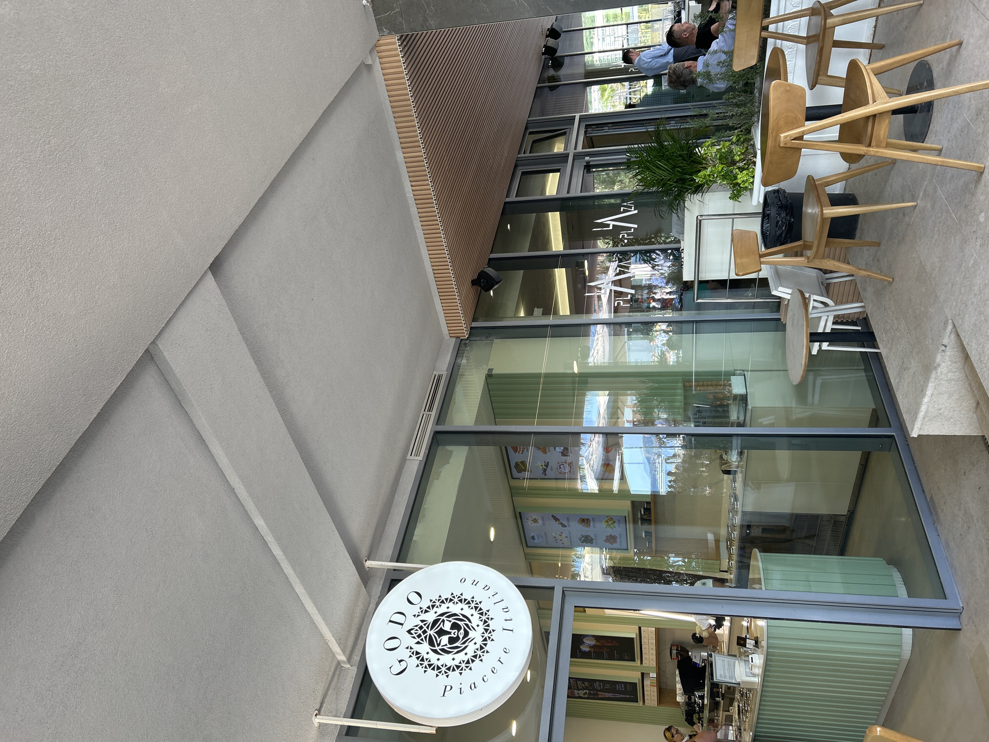 The image depicts the exterior of "Godo Italiano piacere," a business establishment, likely a restaurant or café, situated in Rafailovići, Montenegro. The building features a modern architectural design with large glass windows offering views of the interior and reflecting the outdoor surroundings. A prominent round sign with the business's name in stylized font and a decorative geometric design is affixed to the building's exterior.
Inside the establishment, a counter with a light green, corrugated facade and a cash register can be seen, suggesting a point of service. People are visible inside and outside the building. Outside, wooden chairs and tables are arranged on a patio, where individuals are seated and engaged in conversation or relaxation. The flooring of the outdoor area is paved.
The image is taken from a street-level perspective, showcasing the business's presence within its urban context. The natural light and reflections on the glass create a dynamic visual effect, highlighting both the interior and exterior elements of the scene.
