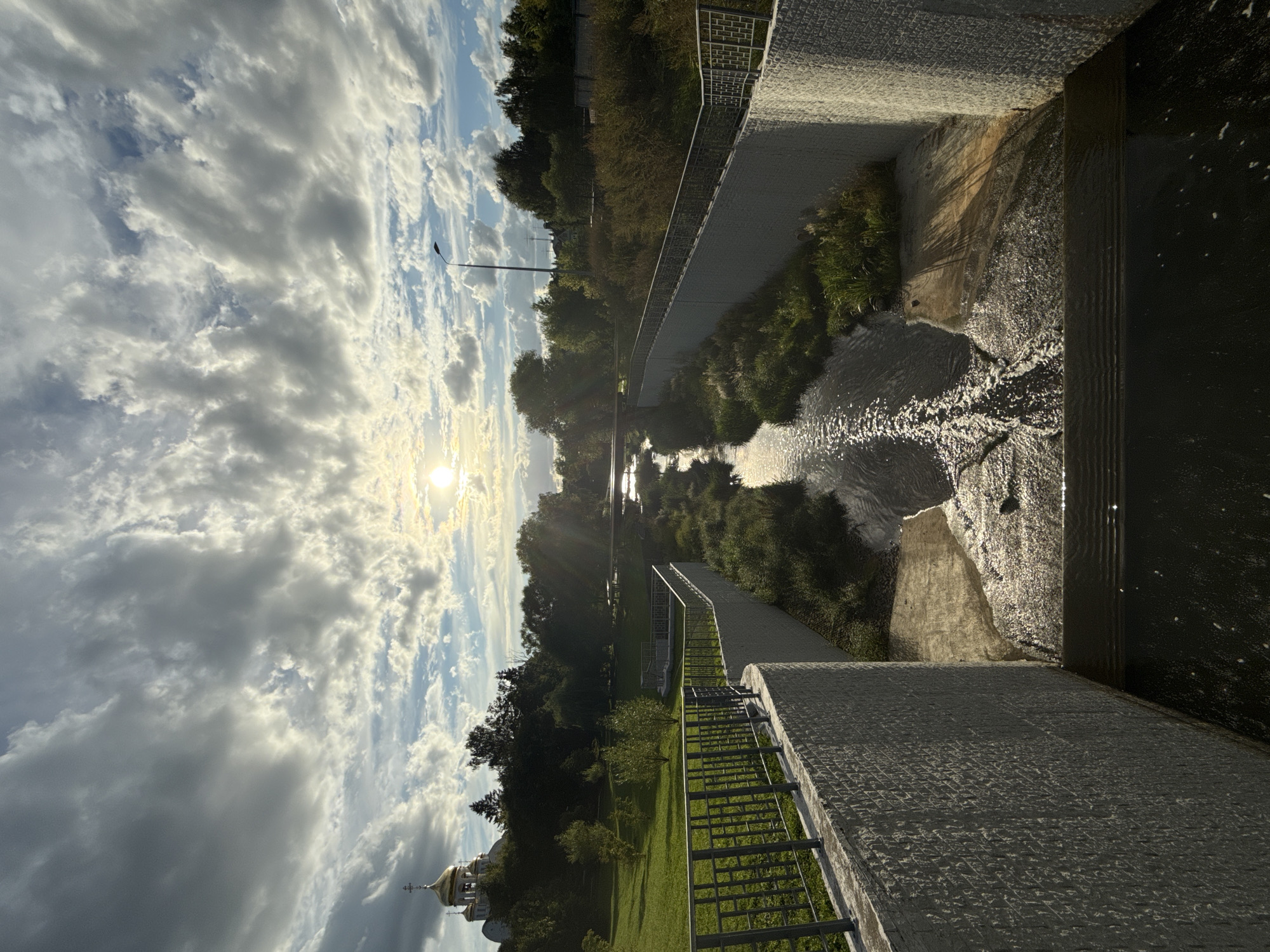 This is a wide-angle, full shot, eye-level, outdoor, vertical photograph of a canal or stream flowing through a city park. The sun is setting and is visible in the upper center of the image, casting rays of light through the clouds and reflecting off the water. The sky is filled with dramatic, wispy clouds.
On the left side of the frame, a golden-domed church or cathedral with multiple spires is visible on a grassy hill. Trees line the horizon behind the church and across the park. The canal is lined on both sides by modern, grey, textured concrete walls. A metal railing runs along the top of the wall on the left. The water in the canal is flowing and appears shallow in parts, with greenery growing along its banks. There are some concrete structures within the canal, creating small waterfalls or steps in the flow of the water. A lamppost is visible on the right side of the frame, behind the trees. The overall scene depicts a serene park setting with a blend of natural elements and urban infrastructure. There are no people or animals visible in the image.