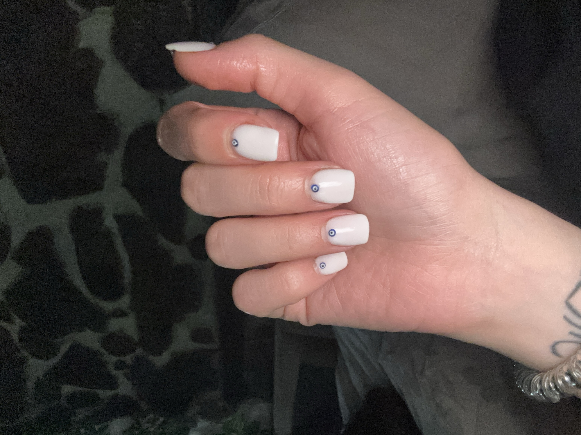 A right hand with white, square-shaped nails is positioned prominently in the center of the frame. Each nail, except the thumb nail, is decorated with a small, blue eye design. The hand is pale with visible skin texture and light reflections, suggesting a slightly oily surface. The hand is in a closed, relaxed position. A metallic bracelet is visible on the wrist. A partial tattoo is observed on the wrist, slightly obscured. The background is dark and out of focus. A stone wall pattern is vaguely visible to the left, and dark grey or black fabric-like material is behind the hand and wrist. There is no indication of the location being Belopoptsi, Bulgaria.