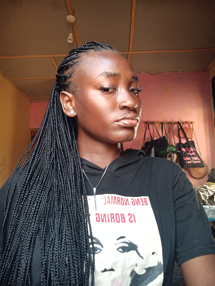 A young woman with long, black braided hair gazes to her right, wearing a black hooded shirt with a print of a person's face and the phrase "Being normal is boring". She has a silver cross necklace. The background features a ceiling with an exposed lightbulb, a pinkish wall, and hanging bags. Her skin appears to have a natural glow, and she wears small stud earrings.