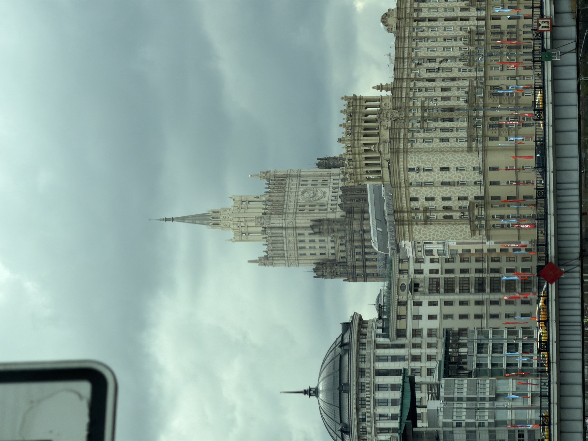 This image captures a vibrant urban scene in Moscow, Russia, depicting a skyline dominated by a striking Stalinist Empire-style skyscraper under an overcast sky. The photograph is taken from an elevated perspective, possibly from a bridge or another building.
In the center, a towering, light-colored building stands out with its intricate, ornate architecture, characteristic of the "Seven Sisters" skyscrapers. It features numerous windows, decorative elements, and culminates in a sharp spire that reaches towards the cloudy sky. A large clock face is visible on one of its lower, prominent towers.
To the right of this central structure, a multi-story building with a more modern, yet still substantial, facade is visible. Its numerous windows reflect the dull light of the day. Adorning the exterior of this building are multiple small, rectangular banners or flags, alternating between blue and red, adding splashes of color to the otherwise muted palette. Visible text on some blue flags reads "УВЕА," and on some red flags, "DOR." A small, red square sign with a white Cyrillic letter "В" is also affixed to this building.
In the left foreground, partially obscured by a blurred object, is another large building featuring a distinctive dark, domed roof with a central finial. Its facade appears to be made of lighter stone with classical architectural elements. The blurry object in the bottom left corner is a white sign with a thick black border. The bottom right shows the edge of a bridge or walkway.
The overall impression is one of grandeur and bustling city life, despite the somber, cloudy weather. The time of day appears to be daytime, though the lack of direct sunlight suggests an overcast day.
