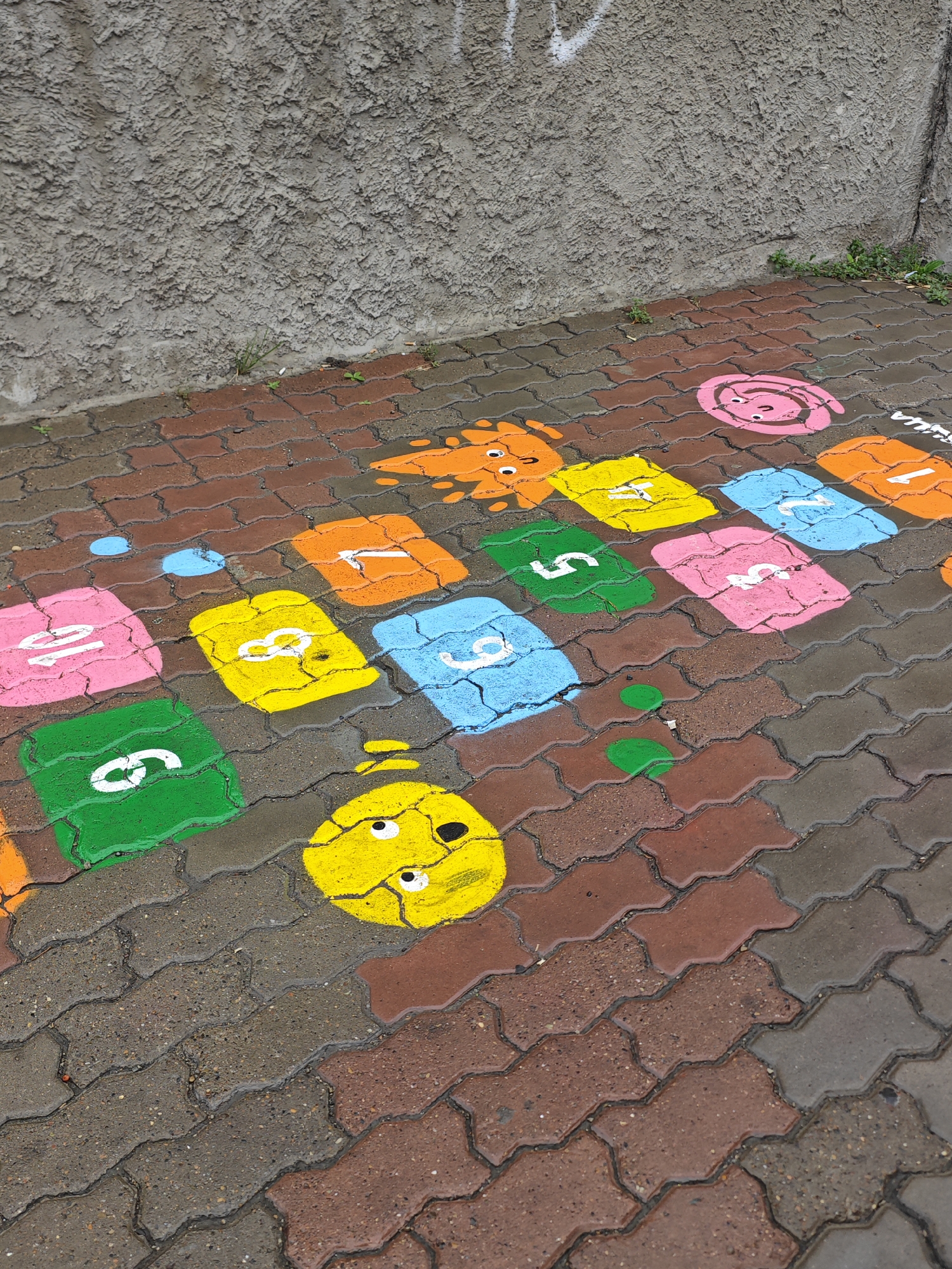 The image shows a wet, textured pavement with colorful painted numbers and cartoon-like figures, resembling a hopscotch or number game, on individual interlocking brick pavers. The colors include orange, yellow, pink, and blue, with white numbers painted on them. Some of the painted figures are round with eyes. The pavement is next to a rough, gray concrete wall, with some small green plants growing near the base of the wall. The scene appears to be an outdoor urban environment. The image is taken from a slightly elevated angle, looking down at the pavement. There are no people visible in the image. The image is not tagged with any specific location in the city of Krasnoyarsk, Russia.