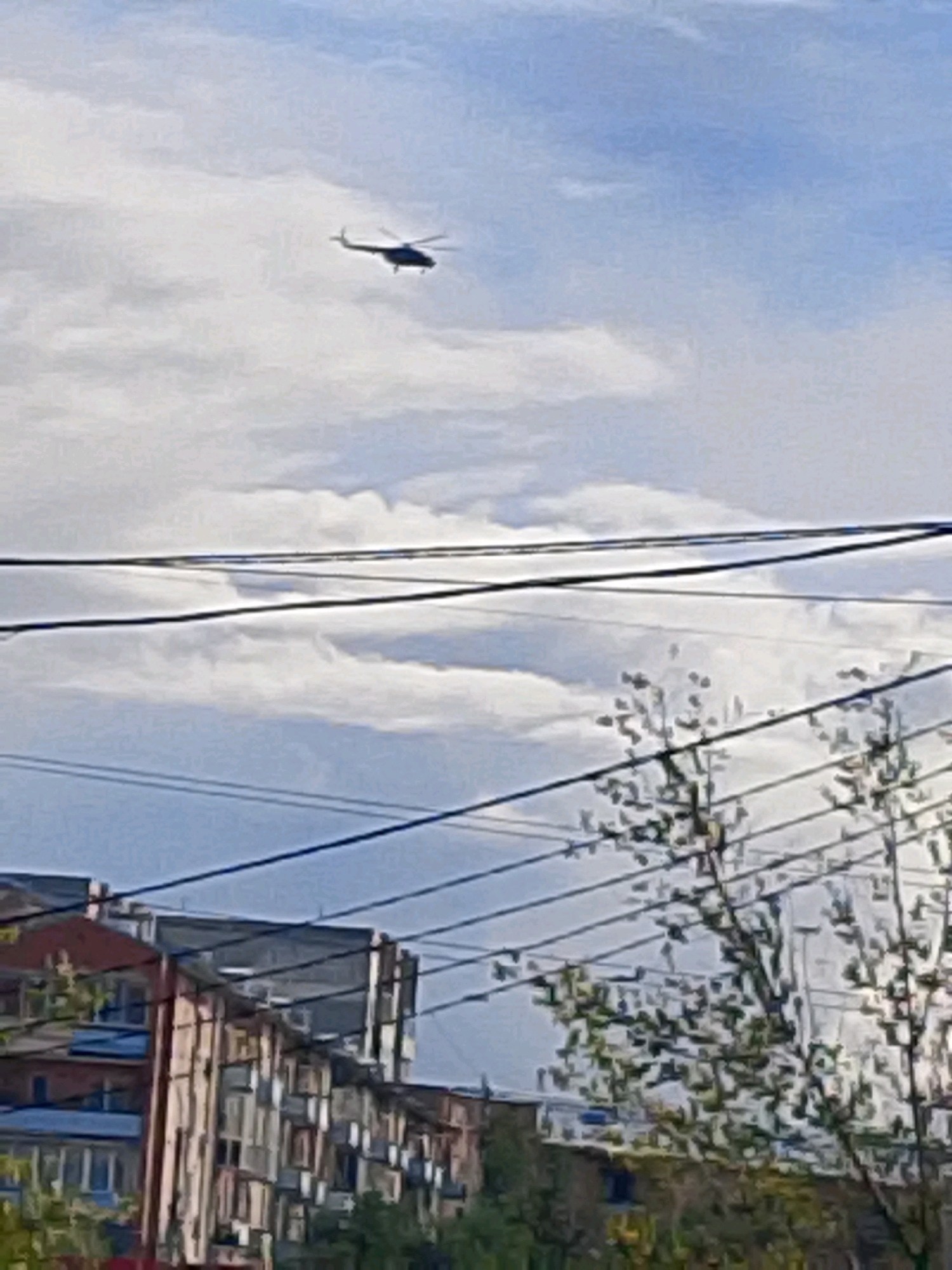 In Kyzyl, Russia, a dark silhouette of a helicopter is captured against a cloudy sky. The helicopter is in flight, angled slightly to the left, with its rotor blades visible. Below the helicopter, power lines crisscross the frame, obscuring parts of the scene. In the lower portion of the image, multi-story buildings with numerous windows are visible, suggesting an urban environment. Trees with sparse foliage are also present in the foreground, with some appearing to be dead or winter-bare. The overall scene depicts a cloudy day with a helicopter in the sky above buildings and power lines.