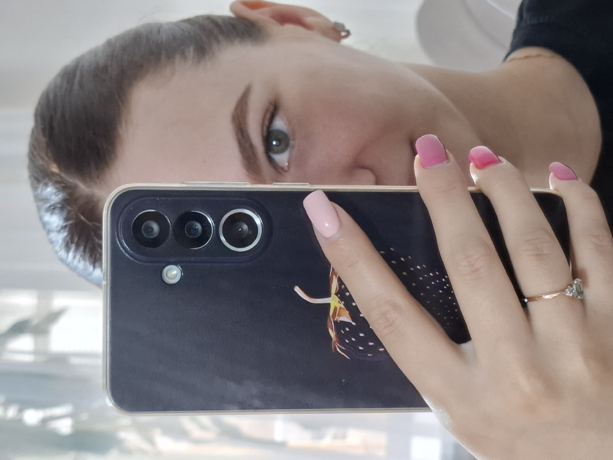 A close-up, inverted mirror selfie captures a young woman holding a smartphone. The woman's face is partially visible on the left side of the frame, showing her forehead, a distinctive green eye, part of her nose and cheek. Her dark hair is pulled back, and she has neatly shaped eyebrows. A small silver stud earring is visible on her right ear.
She holds a dark-colored smartphone, possibly black or dark blue, encased in a clear protective cover. The phone's rear camera module features three lenses stacked vertically with a flash unit beneath them. A faint, abstract design, possibly resembling a spotted feather or insect wing, is visible on the phone's back cover.
Her right hand, holding the phone, is prominent in the foreground. Her fingernails are manicured, with the thumb and index finger painted a light pink, while the middle, ring, and pinky fingers feature a brighter, more vibrant pink polish. A gold-toned ring with a light green or clear central stone, flanked by two smaller clear stones, adorns her ring finger.
The background is softly blurred and light-colored, suggesting an indoor setting with ambient daylight. The overall activity depicts a person taking a self-portrait reflection, with a neutral expression. No text is visible in the image. The setting is an unspecified indoor environment in Chaikovsky, Russia.