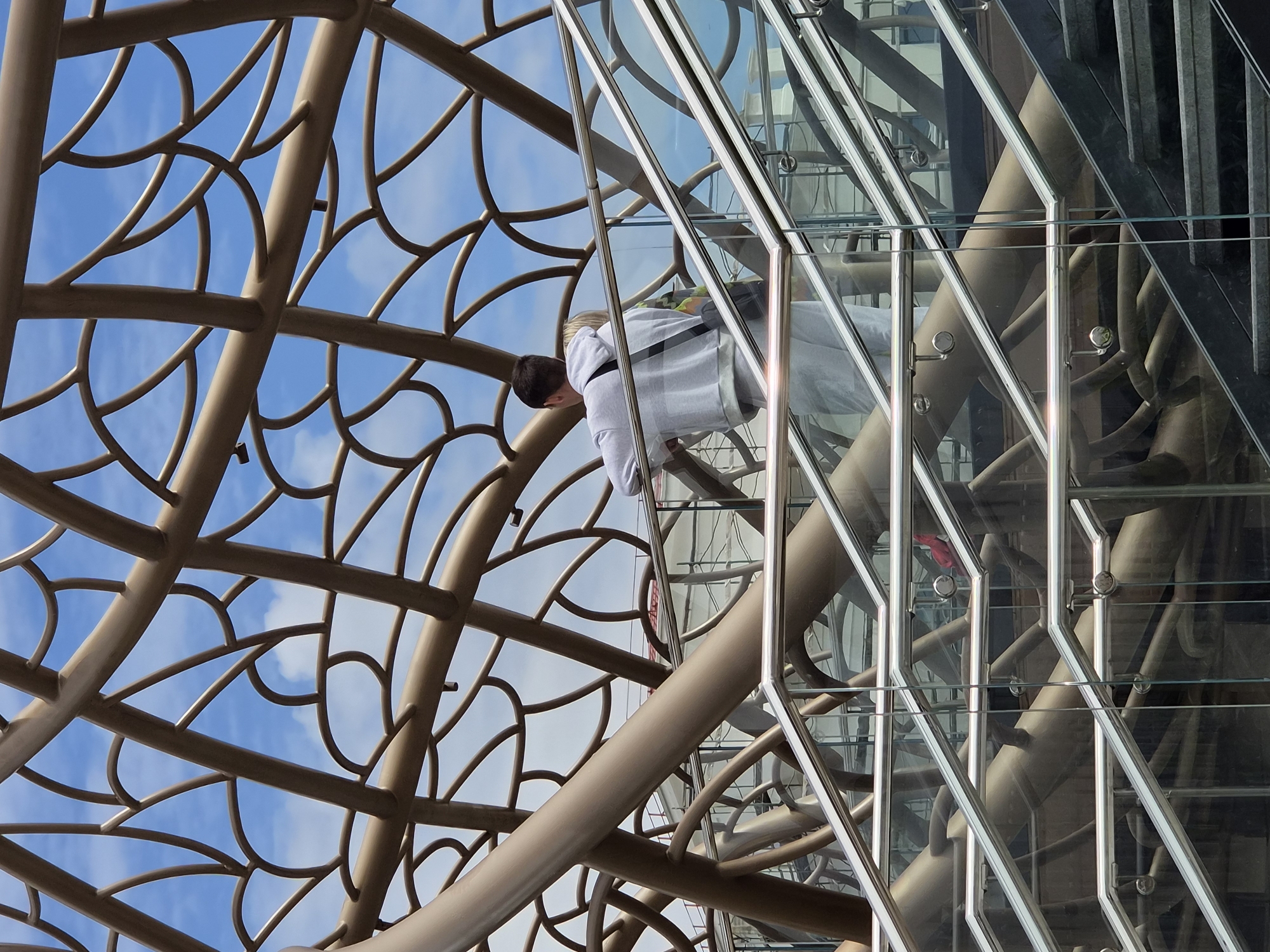 A low-angle, architectural photograph depicts a man standing on a glass-enclosed staircase, with an elaborate network of metal branches or decorative supports visible in the background. This structure is set against a blue sky dotted with wispy clouds.
The man, viewed from the side, has dark hair and wears a white long-sleeved shirt and gray pants. He appears to be looking down, possibly observing something below. The glass staircase is surrounded by metal railings and support structures, creating a grid-like effect.
The metal branches in the background are a light tan or bronze color, and their curved shapes resemble a stylized tree canopy. The photograph's perspective emphasizes the height of the structure and the intricate details of the metalwork and the glass staircase.
This image shows an architectural feature of the Iceberg Skating Palace, a venue in Sochi, Russia, which hosted events during the 2014 Winter Olympics.