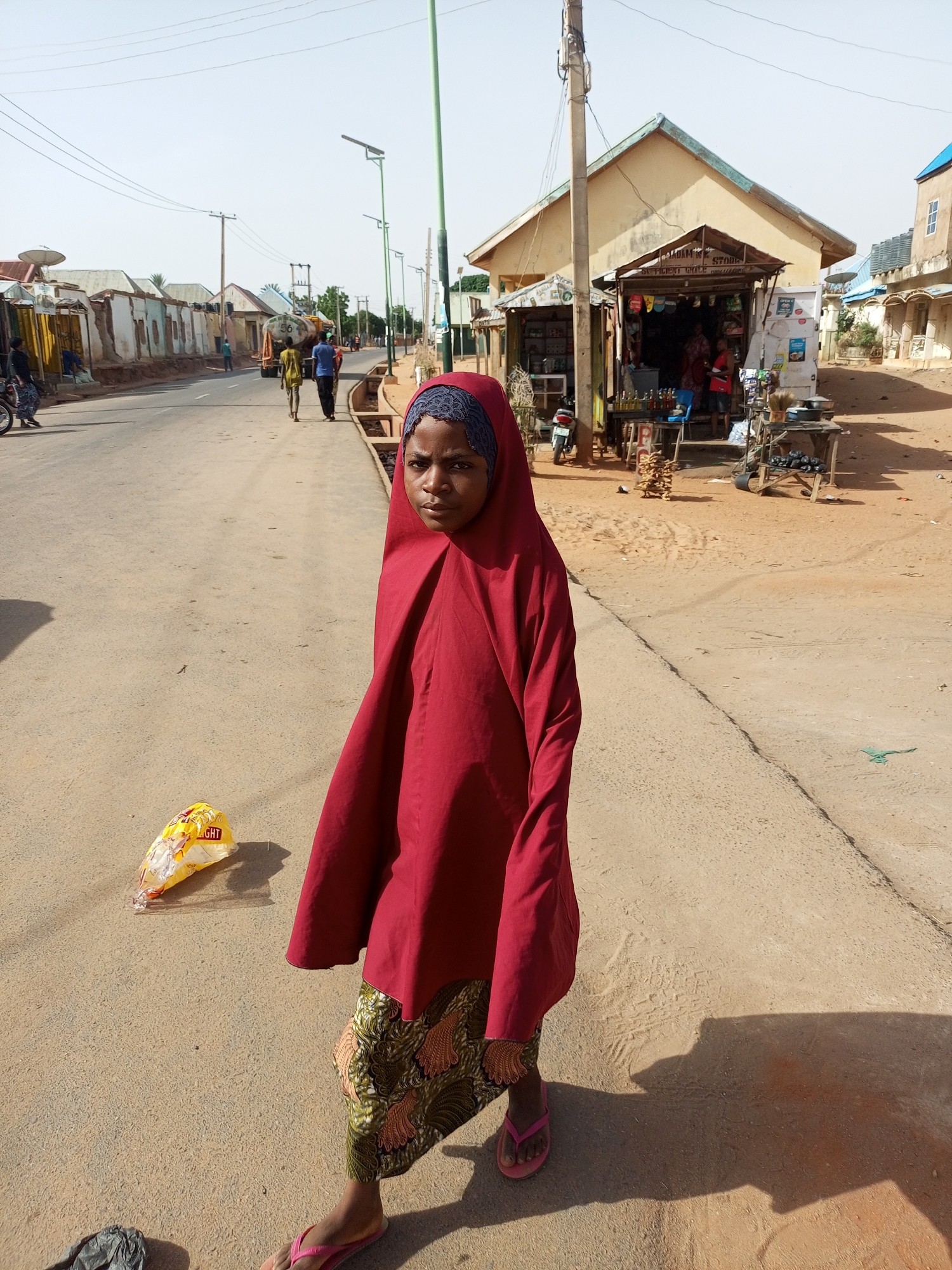 A young girl stands on a paved road in Gombe, Nigeria, wearing a red headscarf and shawl over a patterned skirt, with pink sandals on her feet. Behind her is a small shop selling various items, with people visible inside. The road has several individuals walking in the distance, and buildings line the street. The scene is bathed in daylight. A discarded plastic bag lies near the girl on the road.