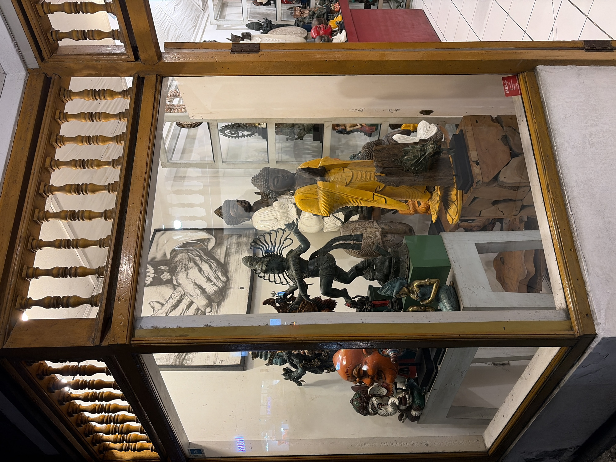This is an outdoor, medium shot of a shop display window in Ubud, Indonesia. The window is framed by weathered, dark brown wooden beams and features a wooden railing at the top. Inside the glass display, numerous statues and sculptures are visible. On the left, a large, orange mask with a wide grin is prominently displayed. To its right, a white statue of Ganesha, the elephant-headed Hindu deity, is partially visible. In the center of the display, a large, yellow-robed statue of Buddha stands with its hands pressed together in prayer. Behind the Buddha, a white statue of Buddha is also visible. Various other statues, including Hindu deities like Shiva and possibly dancers, are arranged on shelves and pedestals. The shop appears to sell artisanal crafts, with a sign for "BAJRA Shop" visible in the lower right corner. The floor outside the shop is tiled, and the overall lighting suggests it is either early morning or late afternoon.