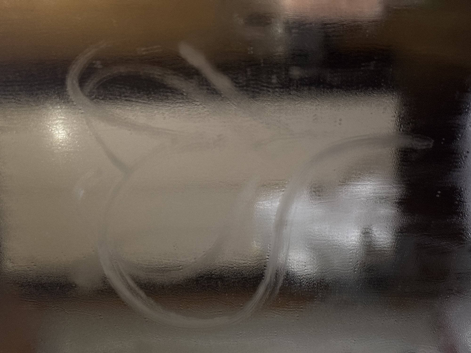 The image shows condensation on a glass surface with circular and curved wiping marks. The background is blurred and indistinct but appears to be an interior scene with varying light and dark areas. There are no identifiable people, objects, or actions in the image that allow for a specific location determination in the city of Chaikovsky, Russia.
