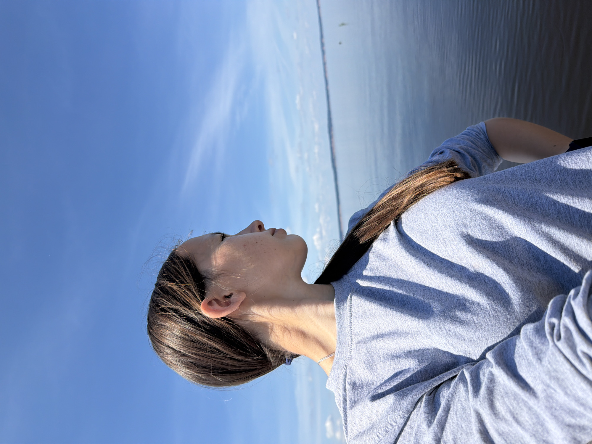 A young woman with dark hair pulled back in a ponytail looks out over a body of water under a clear blue sky with sparse white clouds. She is wearing a grey long-sleeved sweatshirt. The water has gentle ripples and a distant shoreline is visible. The scene appears to be a tranquil outdoor setting, possibly a lake or river, in Stepanovo, Russia.