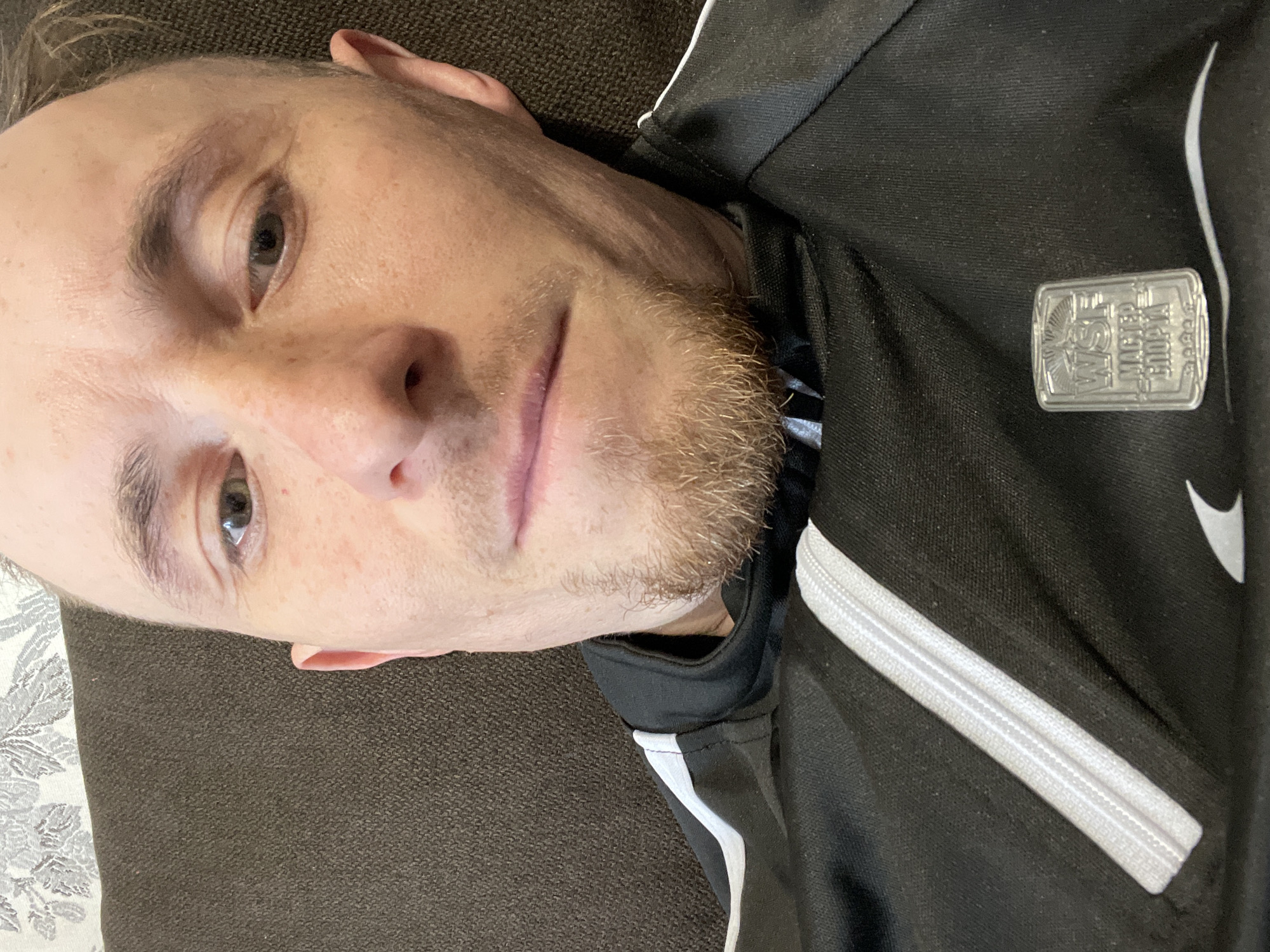 A close-up, slightly angled, indoor shot shows a Caucasian man lying on what appears to be a brown upholstered surface, possibly a couch. The man has light skin, short hair, and a trimmed beard. He is wearing a black jacket with white detailing, including a white Nike swoosh and a rectangular silver-colored pin on the jacket. The pin has embossed writing and imagery.
There is no information to confirm the location to be Sarapul, Russia, nor details about the people, subjects, actions, interactions, or environment.
