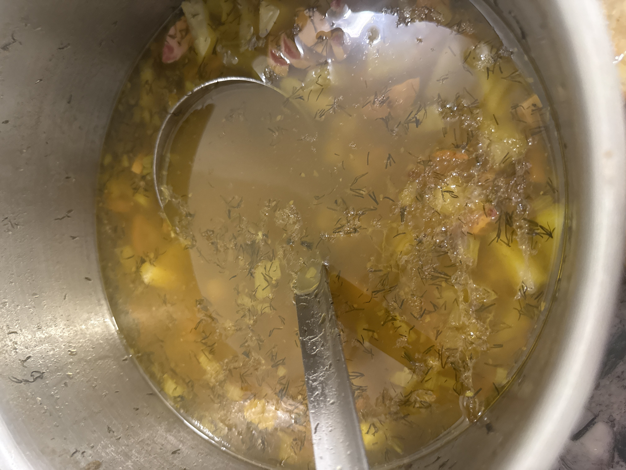 The image shows a close-up, overhead view of a large metal pot filled with a yellowish broth containing pieces of vegetables, herbs, and possibly meat. A silver ladle is partially submerged in the broth. The pot is resting on a surface that appears to be a countertop. There is no indication of the city of Birobidzhan, Russia, or any specific location. The image is a still life focused on the contents of the pot.