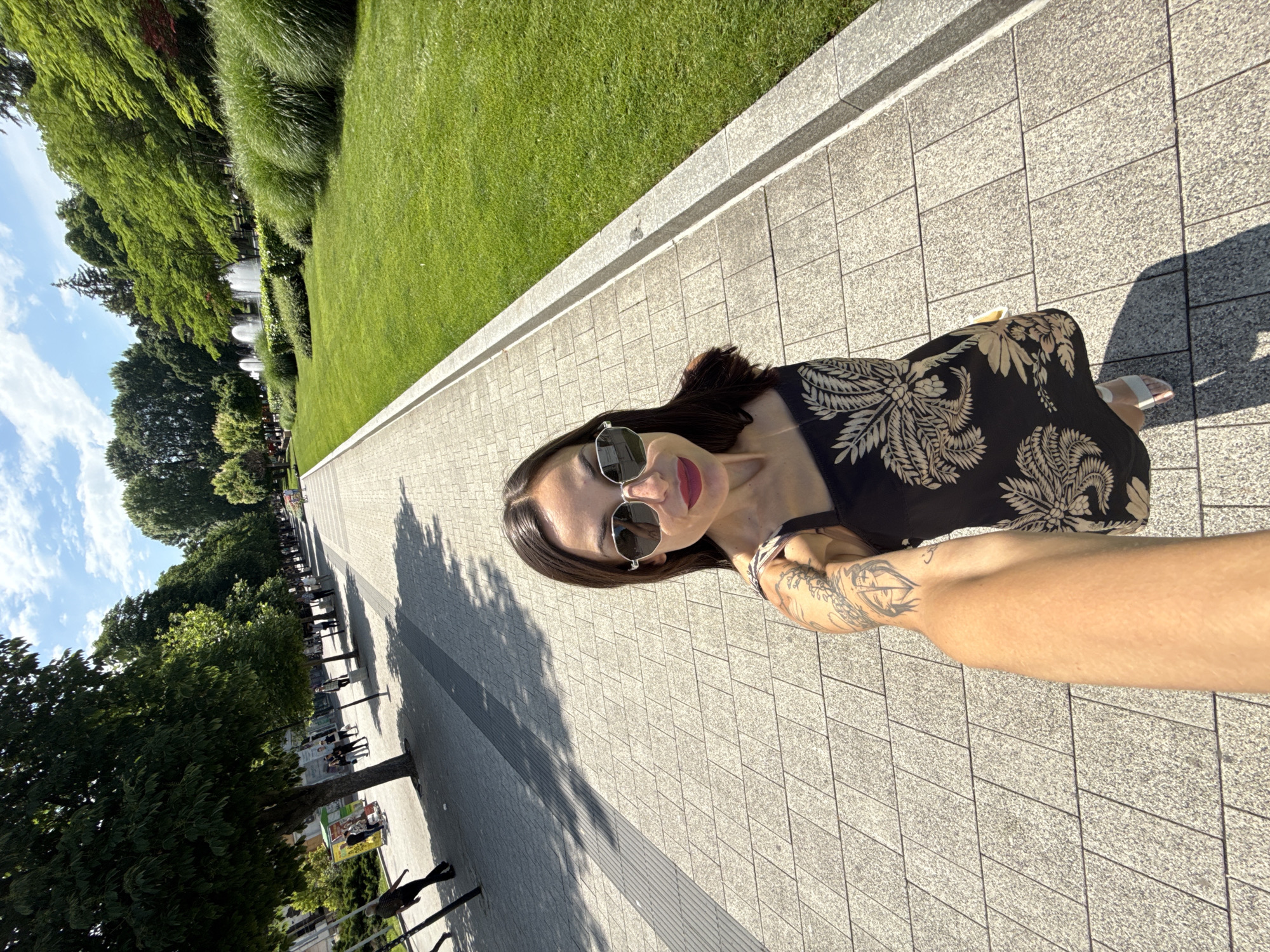 This brightly lit outdoor selfie, rotated 90 degrees clockwise, features a woman captured from the chest up, positioned on the right side of the frame (when upright). She has dark hair, wears sunglasses and red lipstick, and is dressed in a dark garment adorned with a large, light-colored floral or leaf pattern. Her right arm, visible with tattoos, is extended, holding the camera.
She stands on a wide, light-grey paved pathway constructed from rectangular tiles. To her left, a neat strip of green grass is bordered by a low concrete curb. Beyond the grass, a verdant landscape unfolds, showcasing various trees and bushes. In the mid-ground, among the lush greenery, several white water features or fountains are visible. Further along the pathway in the distance, a queue or gathering of people suggests a busy public area.
The image depicts a sunny day, likely midday or early afternoon, given the strong shadows on the pavement and the bright illumination. The sky is partly cloudy with visible blue patches. The scene evokes a pleasant and relaxed atmosphere in what appears to be a public park or garden. No distinct text is legible within the image. This location is contextually identified as Pleven, Bulgaria.