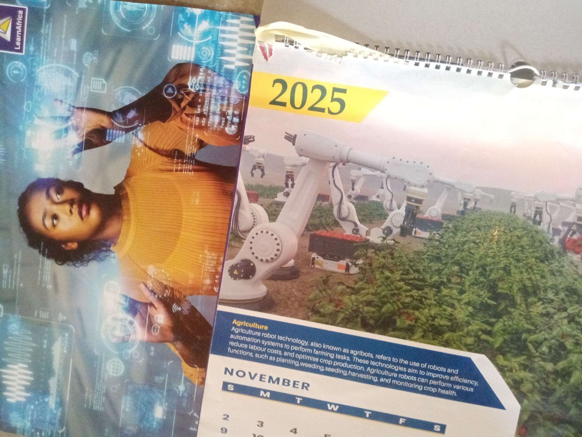 This media file shows a close-up of a calendar page for November 2025, open to a scene featuring agricultural robots in a field of crops. The calendar is part of a larger publication, possibly a calendar or a magazine.
The foreground features a woman, likely a student or professional, interacting with a futuristic holographic interface. She is wearing a yellow top and her expression is focused. The holographic display shows various data visualizations and icons.
The calendar page itself depicts a line of white agricultural robots with articulated arms, positioned among rows of green plants, likely a vegetable farm. Some robots appear to be holding containers. The image suggests a modern and technologically advanced approach to agriculture.
Above the robotic imagery, the year "2025" is prominently displayed in large green numerals on a yellow background.
Below the image of the robots, there is a text box with the heading "Agriculture." This text explains that "Agriculture robot technology, also known as agribots, refers to the use of robots and automation systems to perform farming tasks." It further details how these technologies aim to improve efficiency, reduce labor costs, and optimize crop production, listing functions such as planting, weeding, seeding, harvesting, and monitoring crop health.
The bottom of the calendar page shows the month "NOVEMBER" with the days of the week indicated by their first letters (S, M, T, W, T, F, S). Numbers for the days are partially visible, starting with '2' and '9' in the first column.
The overall impression is one of technological advancement in agriculture, with a focus on innovation and efficiency. The presence of both human and robotic elements suggests a blend of human oversight and automated processes in farming. The location context of Jalingo, Nigeria, implies this is a display relevant to or produced within that region, highlighting the adoption of modern technologies in local agriculture.