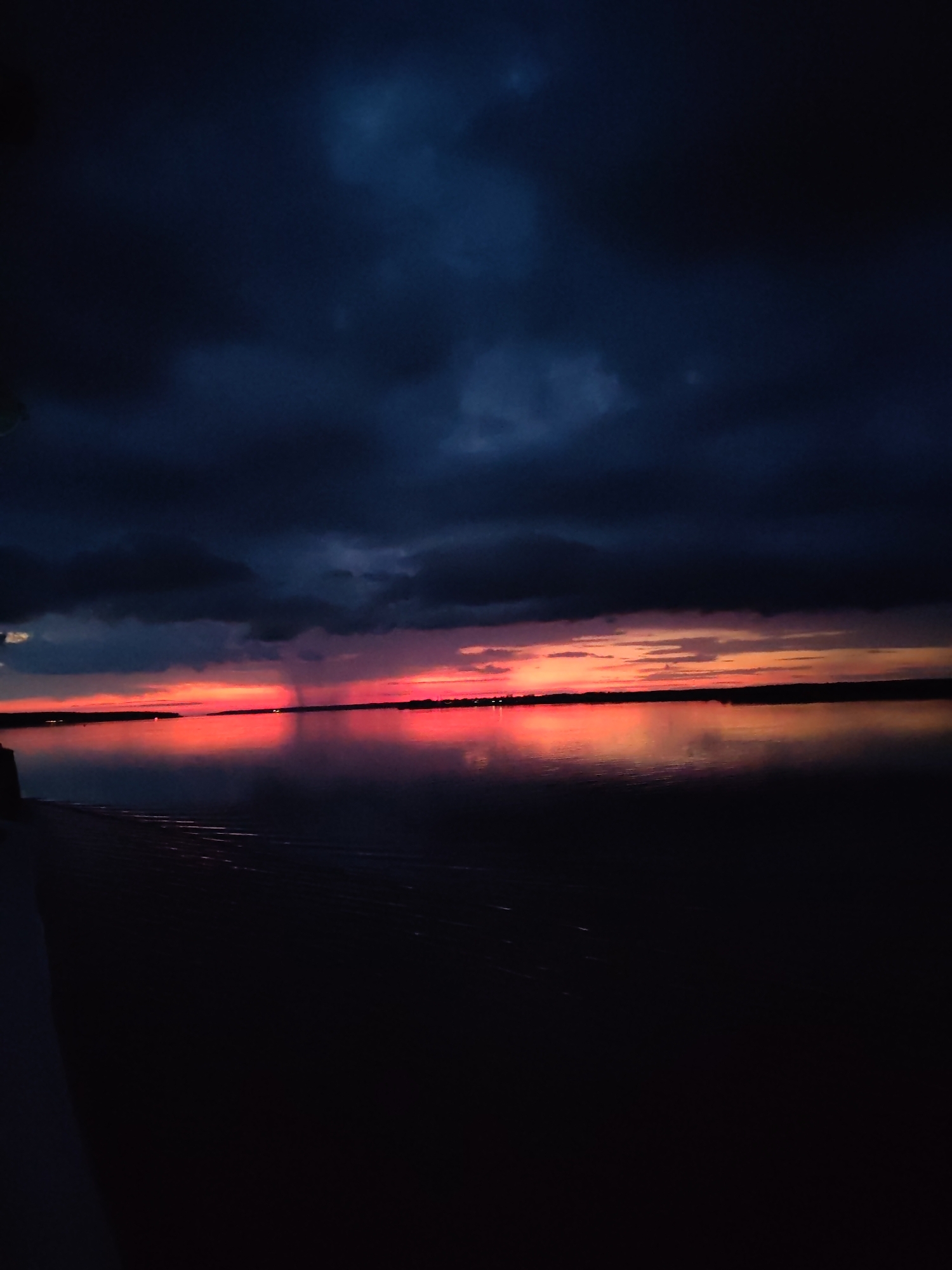 The image depicts a body of water, likely a lake or river, reflecting a dramatic sunset over the city of Bol'shie Mkhi, Russia. Dark, ominous clouds dominate the sky, interspersed with streaks of vibrant orange and pink from the setting sun. The water's surface is smooth, mirroring the fiery sky and the dark silhouettes of distant land and possibly structures. In the foreground, the dark expanse of the water is partially obscured by what appears to be the edge of a pier or embankment. The overall atmosphere is one of twilight and a potential storm, with the last rays of sunlight fighting against the encroaching darkness. No people or specific actions are visible.