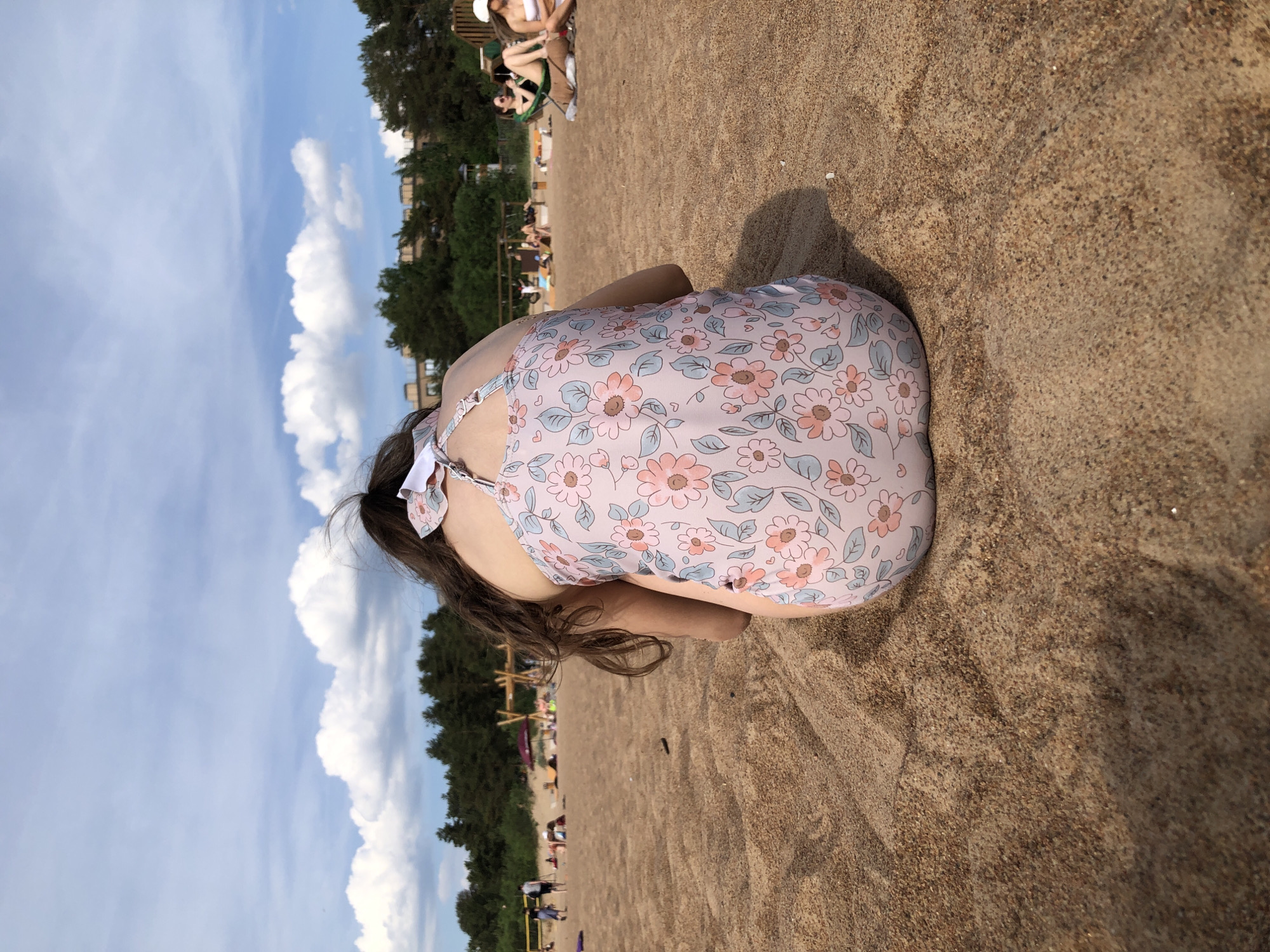 This image depicts a sunny daytime scene on a sandy beach in Sestroretsk, Russia. In the immediate foreground, a young individual is observed from behind, crouching or sitting on the light brown sand. This individual has long, dark hair and is wearing a light pink one-piece swimsuit featuring a repeating floral pattern of pink blossoms and light blue leaves. A white bow is visible at the back of the swimsuit straps.
The midground reveals an expansive sandy beach with visible textures and slight undulations. Several other people are present at varying distances. To the left, two figures stand near the water's edge, and multiple individuals are lounging on the sand. To the right, two people are seated; one is in a green beach chair wearing a dark swimsuit, and another is positioned beside them wearing a white top. A red beach umbrella and several wooden structures, possibly recreational equipment, are also visible.
The background features a dense line of dark green coniferous trees, behind which light-colored multi-story buildings are partially discernible. The sky above is clear and light blue, dotted with scattered white cumulus clouds, indicating fair weather conditions. The natural lighting suggests a mid-day or late afternoon setting, casting subtle shadows on the sand.