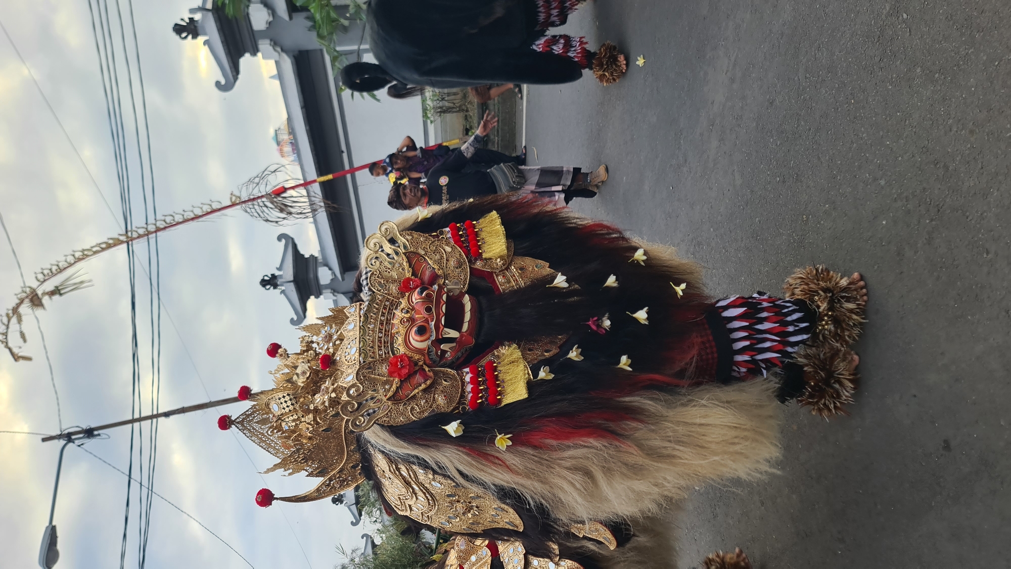 The image depicts a person in traditional Balinese attire, specifically a Barong costume, which is a mythical creature from Balinese mythology. The Barong is characterized by its elaborate, lion-like mask with sharp teeth and piercing eyes, adorned with gold ornamentation, red accents, and red and yellow fringe. The costume itself is voluminous, made of dark fur interspersed with strands of red and blonde hair, decorated with small white flowers. The lower part of the costume features a patterned fabric and furry leg coverings. The person is standing on a paved surface, likely a street, in what appears to be Denpasar, Indonesia.
In the background, other people are visible, some in traditional clothing, participating in what seems to be a procession or cultural event. One person is holding a long pole with a decorative element at the top. The setting includes elements of Balinese architecture, with ornate white structures resembling temple gates or building facades. The sky is overcast, suggesting it might be a cloudy day. The overall mood conveyed by the image is one of cultural vibrancy and tradition. No visible text is present in the image.
