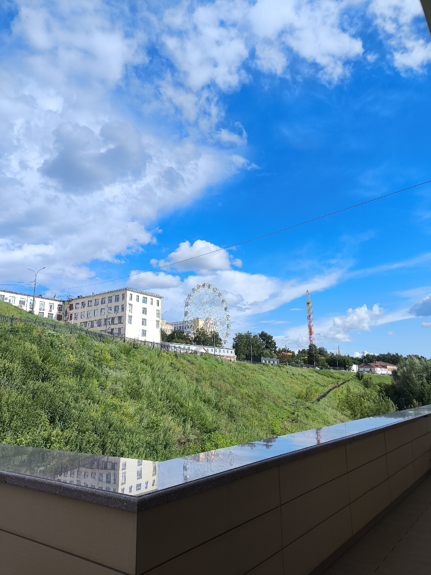 The image depicts a view from a balcony or elevated vantage point overlooking a city park area on a bright, cloudy day. In the foreground, a modern building facade is visible, with a polished reflective surface on top. Beyond this, a steep, grassy incline rises, dotted with greenery. Atop the hill, several buildings are situated, including a large, light-colored apartment complex with multiple windows. To the right of this complex, a prominent Ferris wheel stands tall, and further to the right, a tall, colorful amusement park ride with a thin tower structure is visible. Trees are scattered throughout the scene, and a clear blue sky with scattered white clouds fills the upper portion of the image. The location is identified as a park in the city of Izhevsk, Russia. There are no people or animals visible in the image.