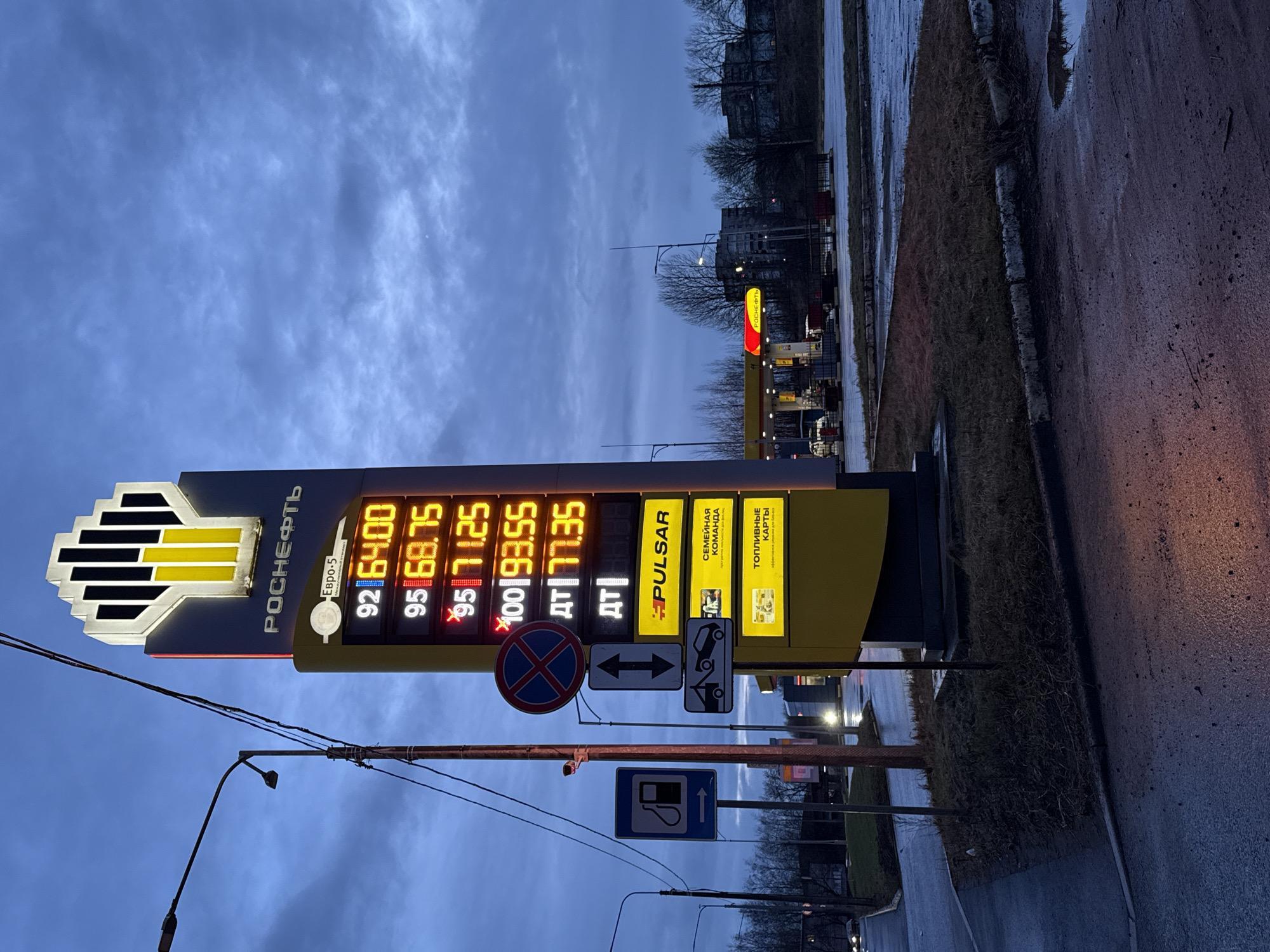 The image shows a Rosneft gas station in what appears to be Saint Petersburg, Russia. A large sign displays the prices for various grades of fuel: 92 octane for 64.00, 95 octane for 68.75, premium 95 octane for 71.25, 100 octane for 93.55, and diesel (DT) for 77.35. The sign also advertises "PULSAR" fuel and "Family Team" fuel cards. Traffic signs are visible, including "No Stopping" and directions indicating possible towing. The environment appears to be overcast with wet roads, suggesting it has recently rained. The location is likely urban, evidenced by the presence of streetlights and buildings in the background.