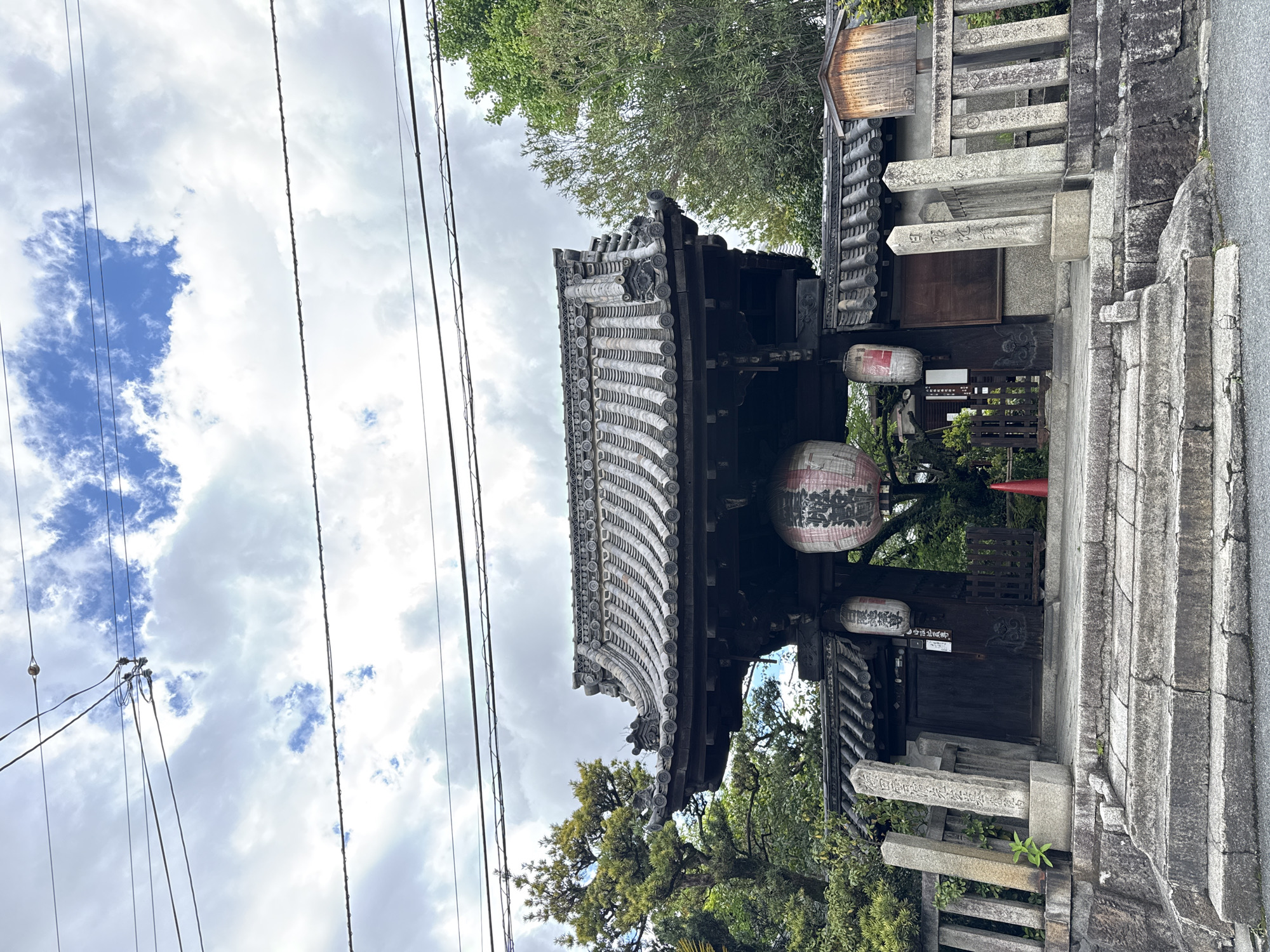 The image depicts the entrance to a Shinto shrine or Buddhist temple in Kyoto, Japan. A traditional Japanese gate structure, composed of dark wood and featuring an elaborately tiled roof, stands prominently. Two paper lanterns with Japanese writing hang from the gate, adding to the cultural ambiance. Adjacent to the gate are stone pillars and steps, suggesting an elevated position or the beginning of a sacred path. Above the gate, a partially visible sky filled with clouds can be seen, along with power lines. The surrounding environment includes trees and greenery, indicating the presence of a garden or natural area.