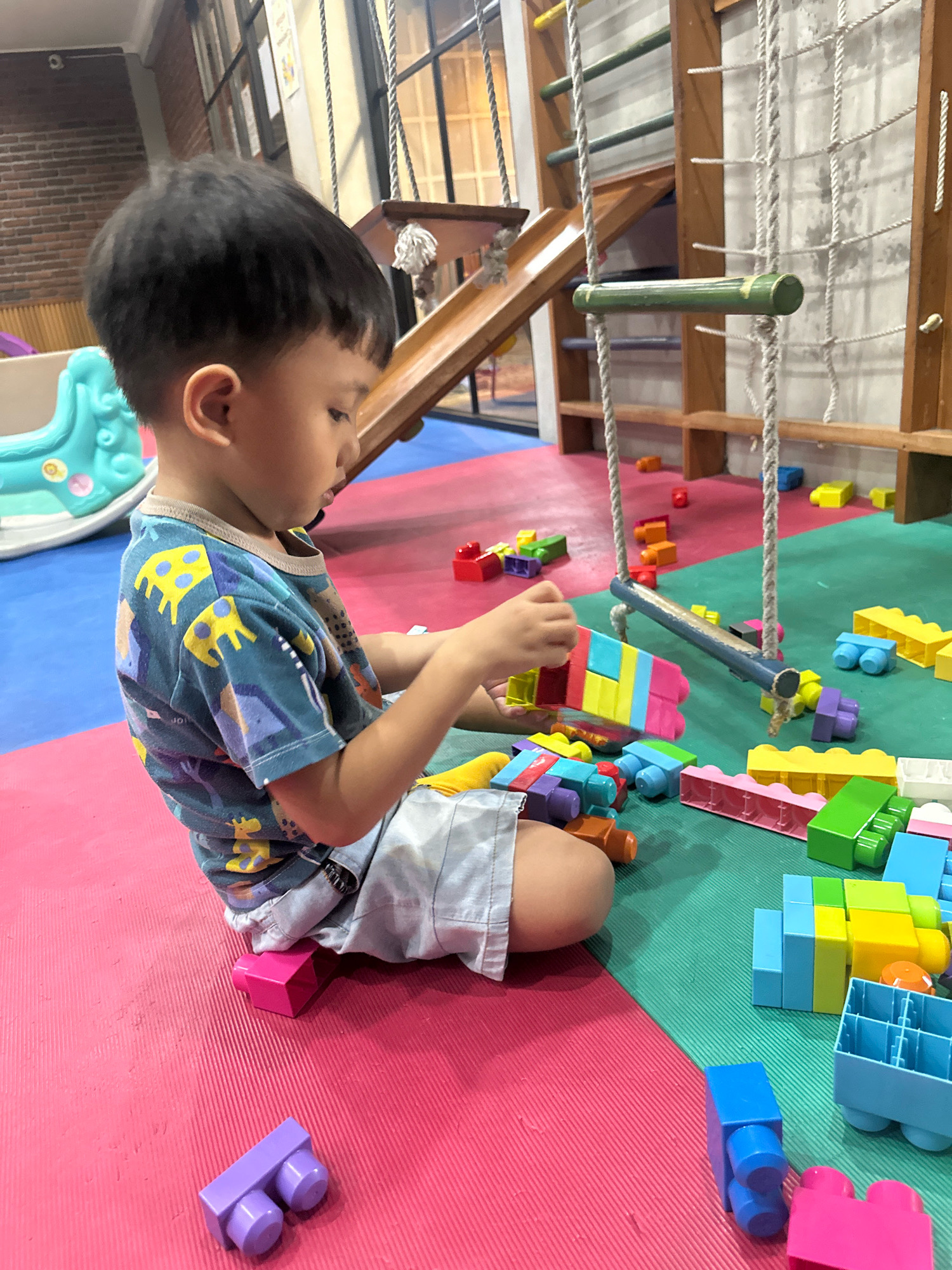 The image shows a young boy sitting on a multi-colored play mat in what appears to be an indoor playground or daycare facility. The boy is wearing a blue t-shirt with a yellow giraffe pattern and light-colored shorts. He is focused on playing with a collection of colorful building blocks, predominantly in red, yellow, blue, green, and pink. Some of the blocks are connected, suggesting he is in the process of building something.
The environment features a wooden climbing structure in the background with a slide, ropes, and horizontal bars. The floor is covered with interlocking play mats of pink, blue, and green. Other children's toys, including a turquoise plastic toy, are visible in the background. The lighting suggests an indoor setting with natural light from windows or a well-lit interior. There are no explicit indicators of the specific location in Mojokerto, Indonesia, beyond the general context of a children's play area.