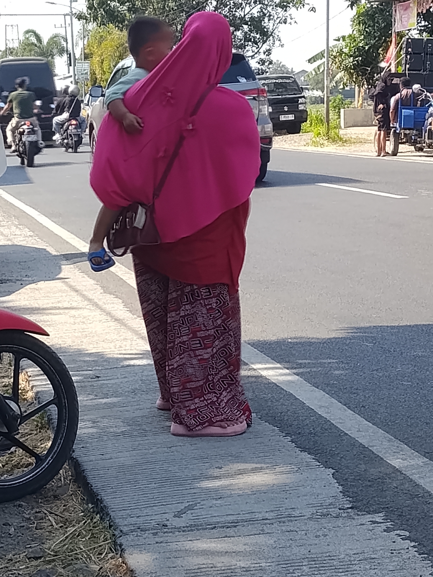 The image depicts a scene on a paved road in Buduan, Indonesia. A woman, from the rear, is carrying a child on her left side. She is wearing a bright pink headscarf and a red garment with a patterned skirt. She is also wearing blue sandals on her left foot and pink sandals on her right foot. The child is wearing a light blue shirt. They are standing on a concrete sidewalk next to the road. On the road, there are several vehicles, including a black car and multiple motorcycles. In the background, there are trees and buildings. The lighting suggests it is daytime, with sunlight casting shadows.