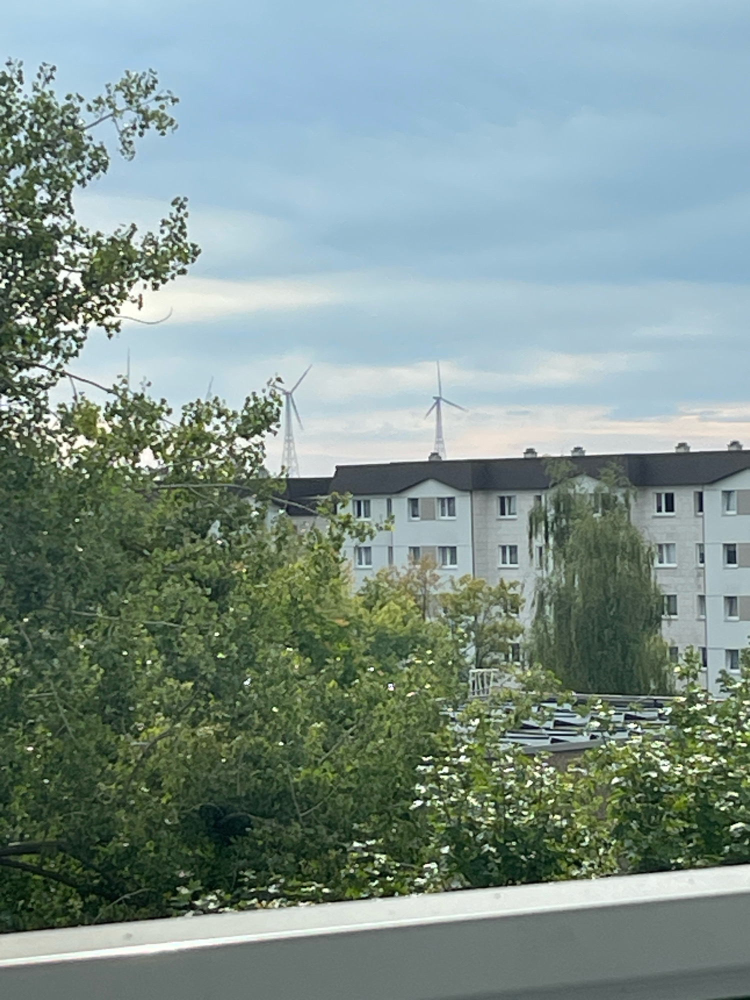 The image shows an outdoor scene with a cloudy sky. In the foreground, lush green trees with some white flowers are visible. Behind the trees, a multi-story apartment building with numerous windows is prominent. Two wind turbines are visible in the distance, one on the left and another on the right, extending above the roofline of the building. The scene appears to be taken from a slightly elevated perspective, looking over the trees towards the building and turbines. The overall environment suggests an urban or suburban setting in Spremberg, Germany.