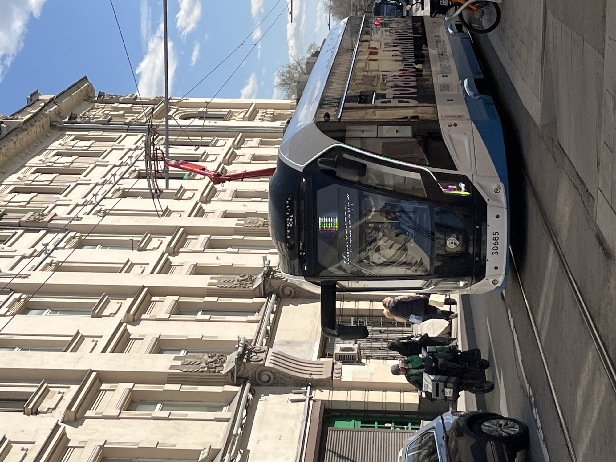 The image depicts a tram traveling on a street in Moscow, Russia. The tram is white with blue accents and has the number 30685 on its front. It is moving along tram tracks that run parallel to the sidewalk. Several people are visible on the sidewalk, including a courier on a motorcycle. A car is parked nearby. The building behind the tram is a multi-story structure with a beige facade. Overhead power lines for the tram are visible against a partly cloudy sky.