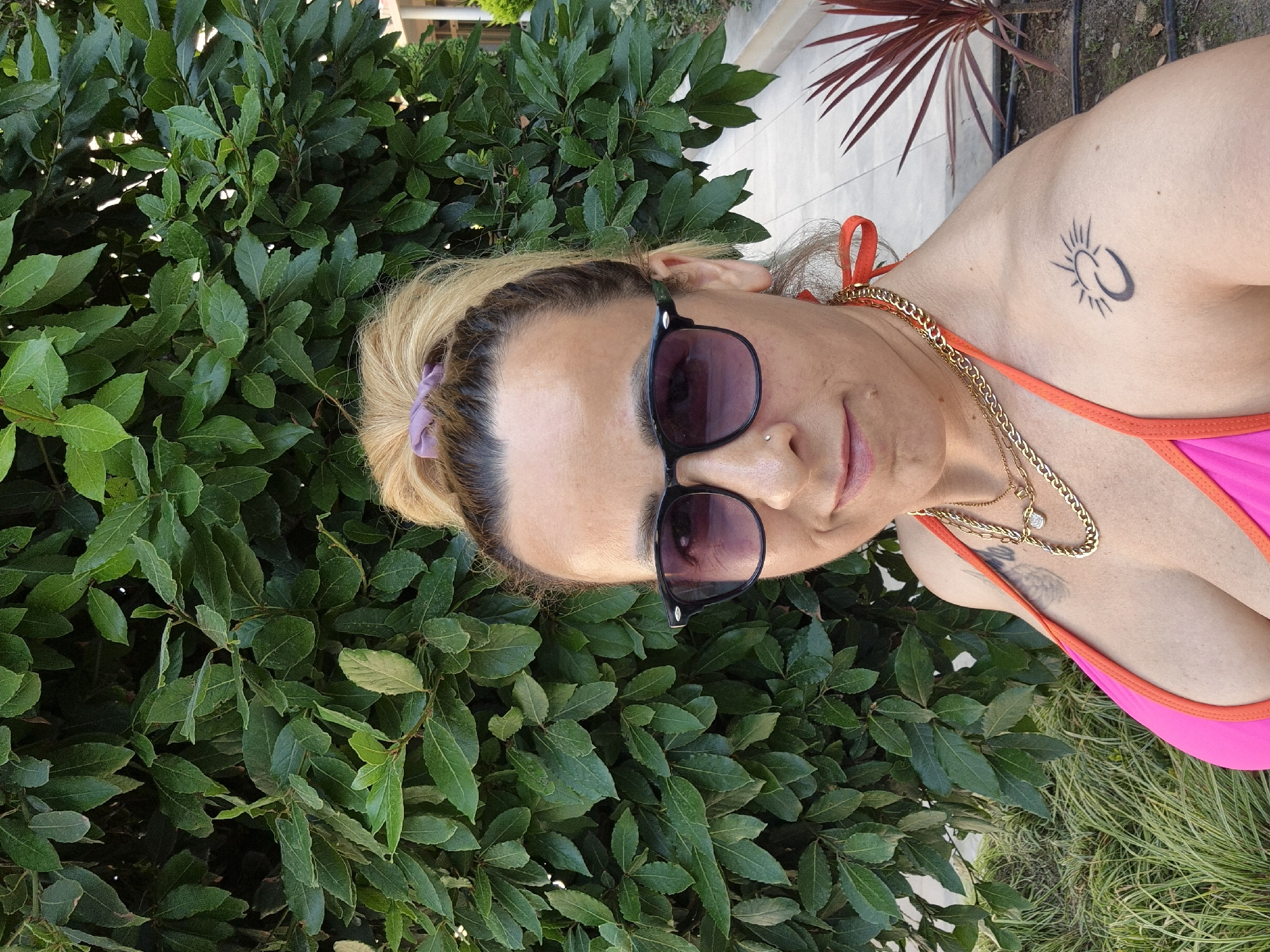 A woman with blonde hair pulled into a messy bun and wearing sunglasses, a pink bikini top, and gold necklaces poses for a selfie. She has a small nose ring. She is standing outdoors in front of a dense green hedge, with a paved walkway and some red-leafed plants visible in the background. A tattoo of a sun with rays and a crescent moon is visible on her left shoulder.