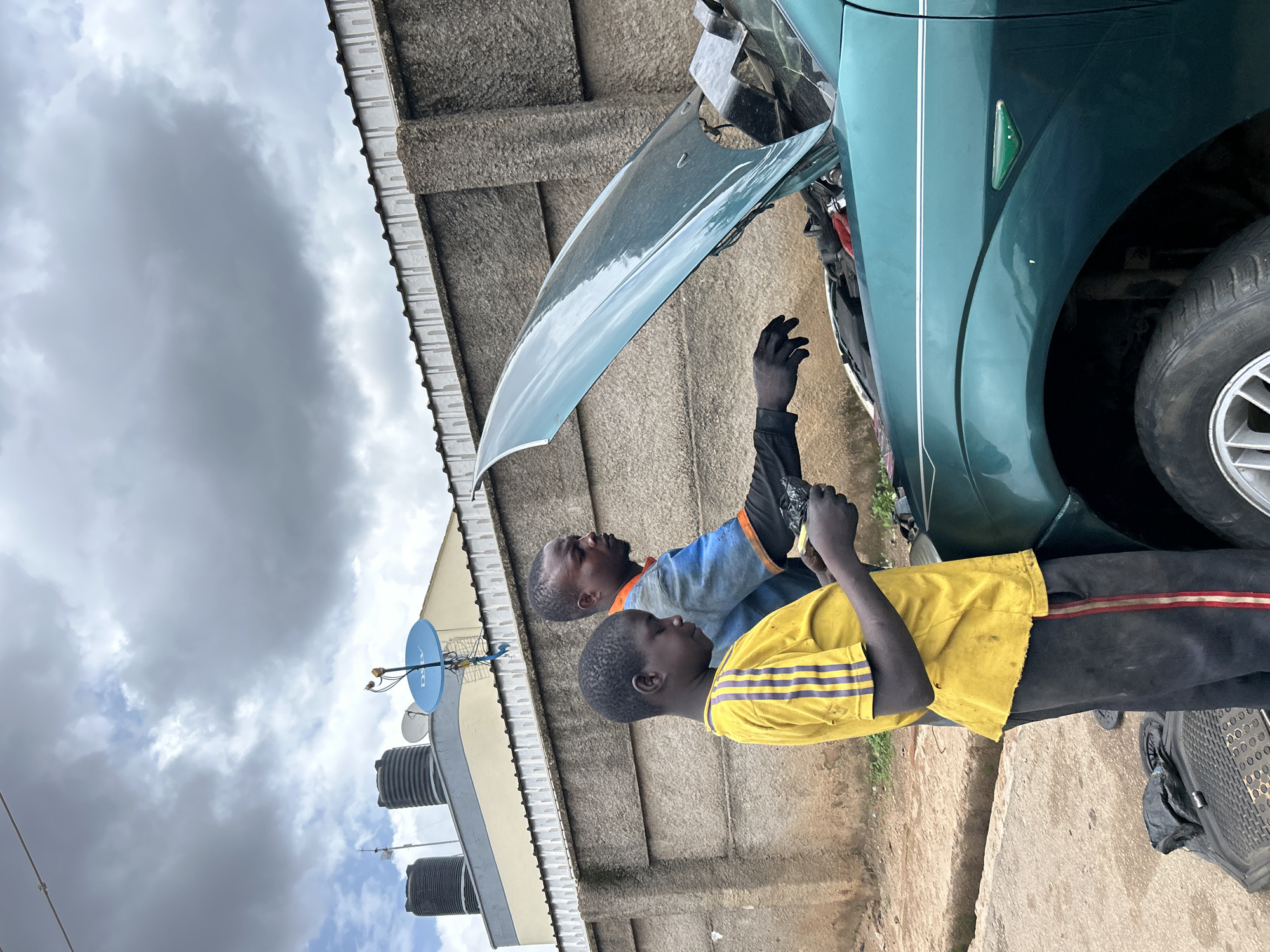 The image depicts two young males working on a green car with its hood open. One individual, wearing a blue and orange shirt, appears to be an older brother or mentor, while the other, in a yellow shirt with blue stripes, seems to be assisting. Both are engaged in mechanical work.
The scene is outdoors, with a textured concrete wall in the background. Above, a cloudy, overcast sky suggests a potentially humid or rainy day, typical for Jos, Nigeria. Visible on the wall are water tanks and satellite dishes, common features in residential areas of Nigeria, further confirming the location context.
The clothing of the individuals suggests a casual, possibly working-class environment. The overall mood appears focused and task-oriented, with no overt display of emotion. The car itself is a mid-sized sedan, with its front-right wheel and fender prominently visible. The ground is a mix of dirt and concrete. There is no visible text in the image. The time of day is indeterminate due to the overcast sky, but it appears to be daytime.