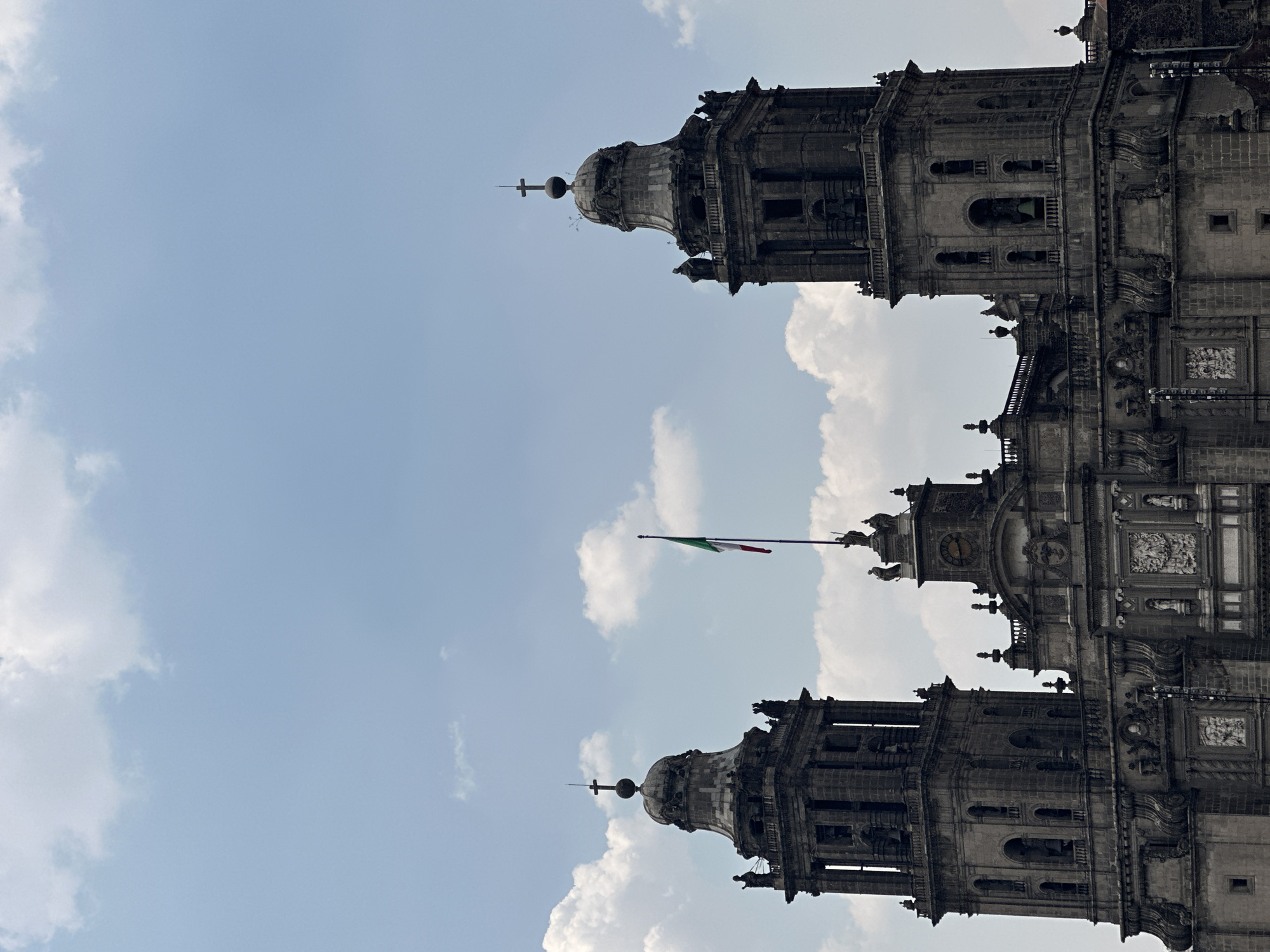The image features the Metropolitan Cathedral of the Assumption of the Most Blessed Virgin Mary into Heaven, located in Mexico City, Mexico. The cathedral's architectural details are visible, including its towers, domes, and façade elements. The Mexican flag is flying atop the structure. The sky is partially cloudy with a blue hue. The image is oriented vertically.
