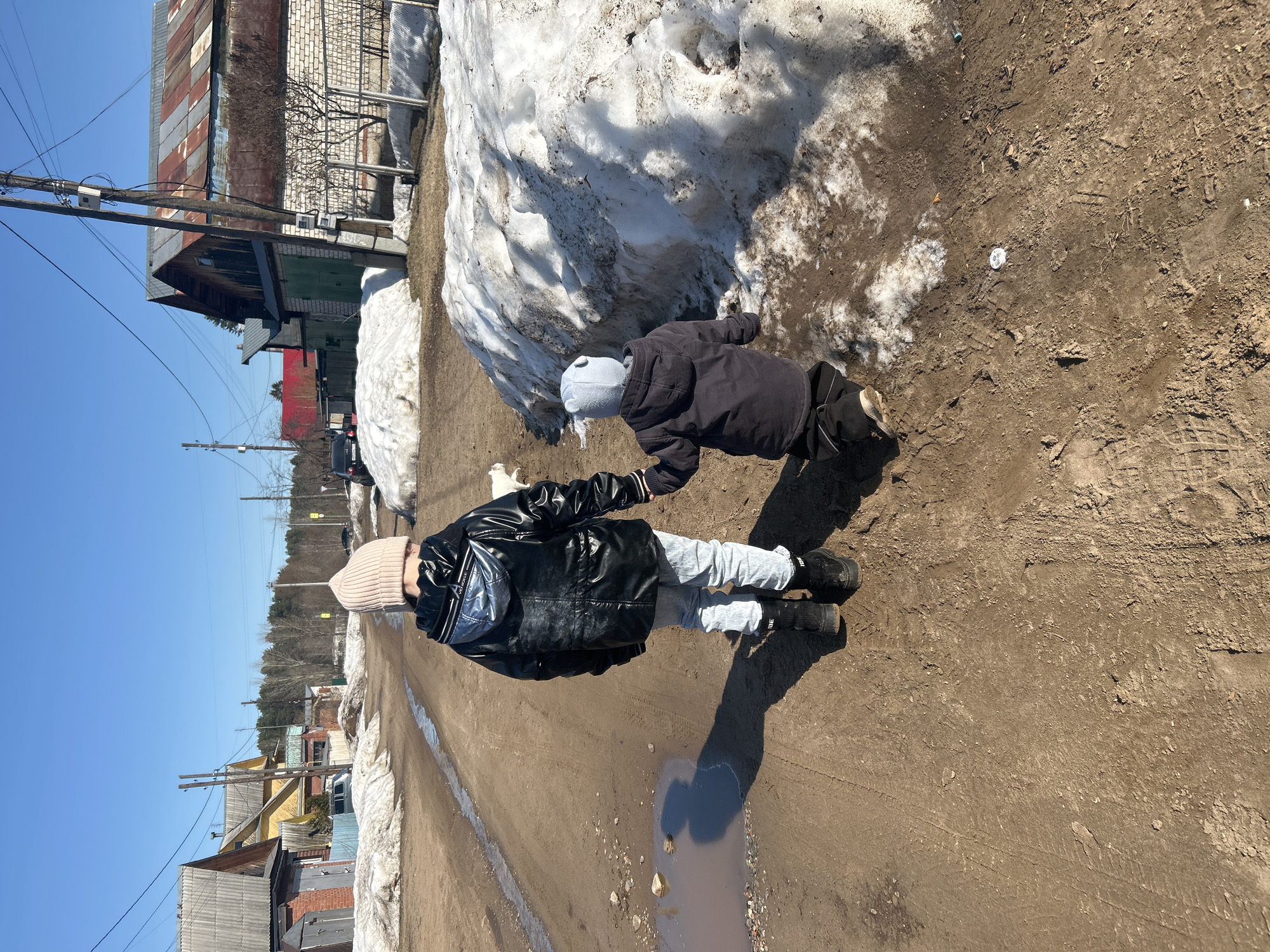 Two people are walking along a dirt road in what appears to be a rural area. One person, presumably a child, is wearing a black jacket, gray pants, a gray hat, and black shoes. The other person, presumably an adult, is wearing a black leather jacket, light blue jeans, a pink beanie, and black boots. They are holding hands. Puddles of water are visible on the road, and melting snow piles are on the side of the road. Buildings and telephone poles line the road. The sky is clear and blue. Based on the style of the buildings and the remnants of snow, the location is likely in Chaikovsky, Russia, as requested.