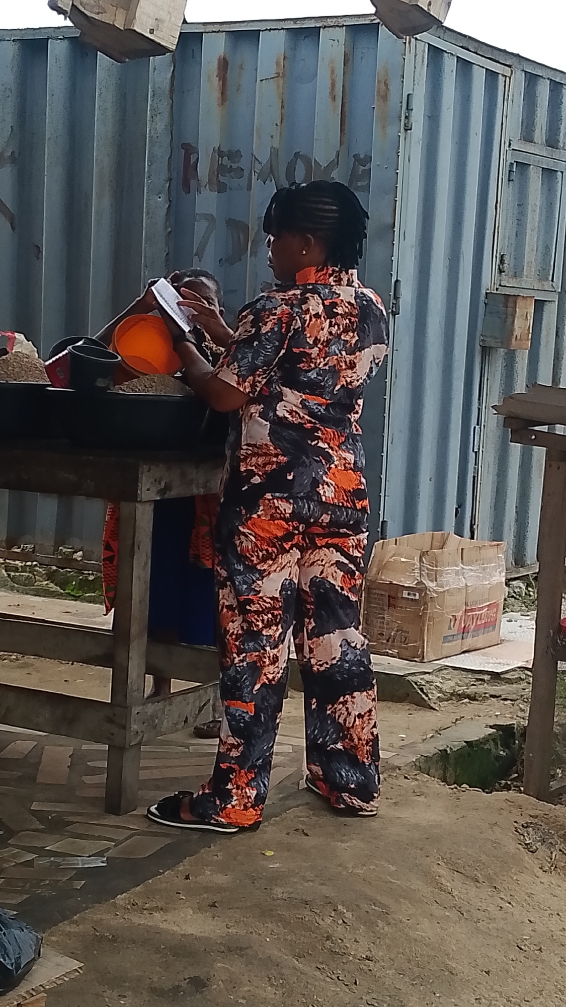 In Warri, Nigeria, a woman with braided hair stands at a wooden table, wearing a vibrant orange, black, and white patterned shirt and matching pants. She is holding an orange bowl and a notebook, appearing to be either organizing or accounting for items on the table. To her left, another person is partially visible, also interacting with the items on the table. Behind them, a large, weathered metal container with faded red lettering, likely a shipping container, serves as the backdrop. A discarded cardboard box sits on the ground to the right of the scene. The ground beneath them is paved with unevenly laid tiles, and the surrounding area appears to be an outdoor market or storage space.