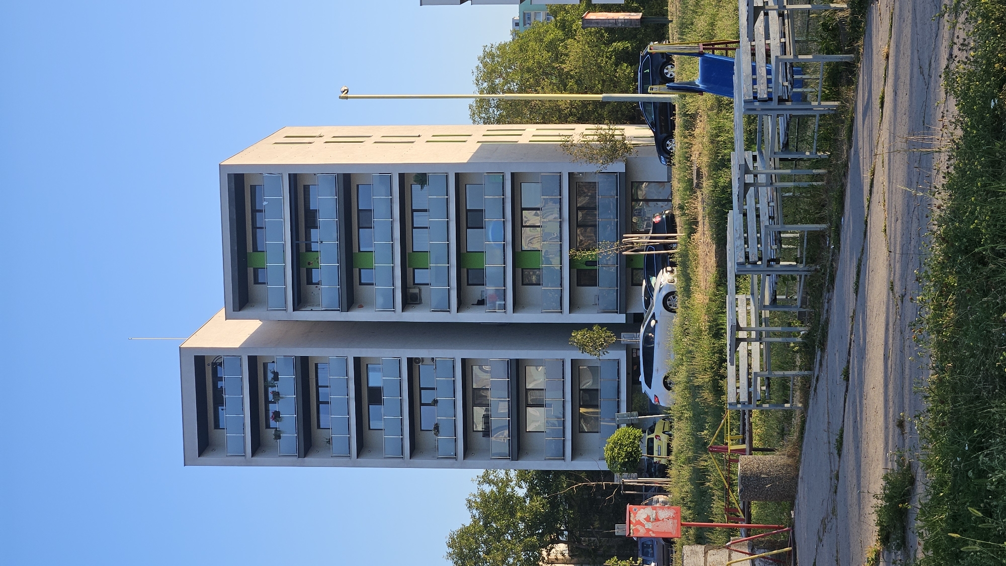 The image shows two modern, multi-story apartment buildings in Prešov, Slovakia, against a clear blue sky. The buildings are constructed with concrete and glass balconies, with some green accents on the upper floors. Several cars are parked along the street in front of the buildings, and trees line the perimeter. In the foreground, there is a paved area with picnic tables and benches, and a small blue slide. The scene appears to be a residential area on a sunny day.