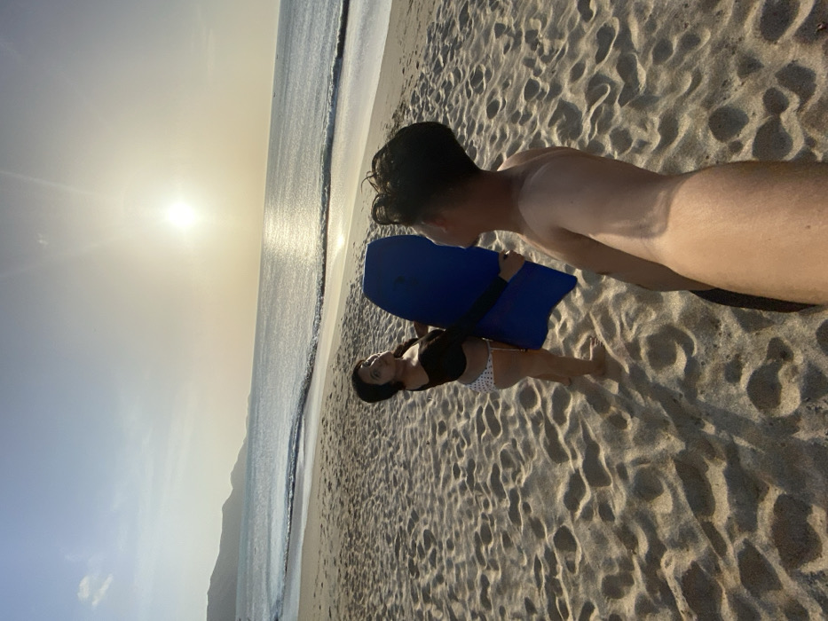 The image depicts two individuals on a sandy beach at what appears to be sunset or sunrise in Juan Griego, Venezuela. A female subject stands on the left, facing partially towards the camera. She wears a black long-sleeved crop top and white polka-dotted bikini bottoms. Her left arm supports a blue bodyboard positioned vertically in front of her. To the right, partially visible in the foreground, is a male subject, shirtless, with dark hair, whose right arm extends towards the camera, suggesting he is taking the photograph. The beach sand shows footprints and ripples. The calm ocean stretches into the background with small waves breaking near the shore. The sun is low on the horizon, creating a bright reflection on the water. Distant mountains or landmasses are visible on the left side of the horizon. The sky is clear.