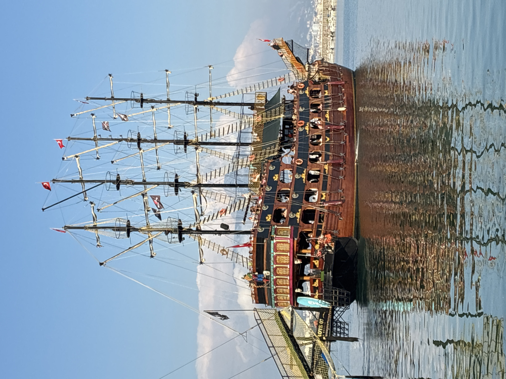 A large, wooden pirate-themed ship is docked in the harbor of Alanya, Turkey, with a clear blue sky and some clouds overhead. The ship has multiple masts with sails furled, rigging, and several decks filled with people. The hull is elaborately decorated with gold trim and colorful accents. There is a ramp leading from a pier to the ship, with a sign that reads "TOYS 07". The ship's reflection is visible in the calm water. In the background, other boats and the cityscape of Alanya can be seen.
