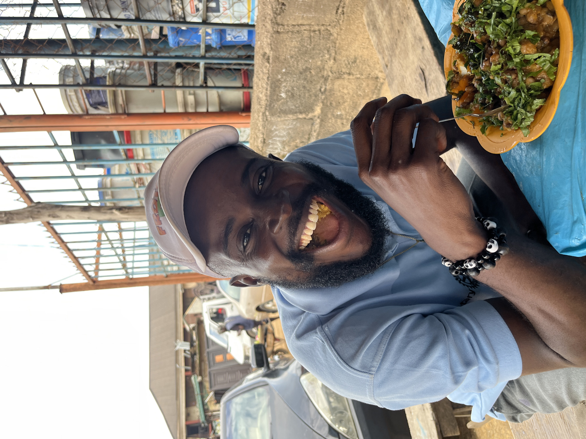 In Gura Top, Nigeria, a Black man with a beard wears a light blue polo shirt and a white baseball cap. He is seated outdoors at a wooden table covered with a blue plastic cloth, holding a fork and eating from an orange bowl filled with food. The food appears to be a stew or bean dish topped with chopped green herbs. He is looking directly at the camera with a wide, joyful smile, showing his teeth. Behind him, a metal fence with orange and blue bars is visible, with stacks of large white and grey buckets behind the fence. To the left of the man, cars are parked, and buildings with brown roofs can be seen in the background. The overall setting suggests an outdoor eatery or market environment.