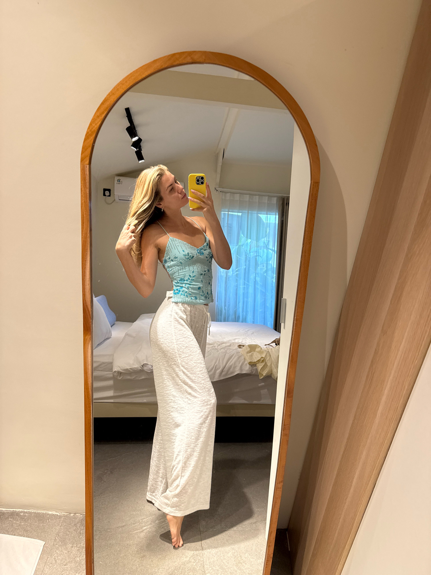 A young woman with blonde hair is taking a mirror selfie in a bedroom. She is wearing a light blue sequined camisole and white wide-leg pants. She is holding her phone in her right hand, which is yellow. Her left hand is touching her hair. In the mirror, a bed with white linens and blue pillows is visible. Behind the bed, sheer white curtains cover a window. The room has light-colored walls and a tiled floor. A wooden arch-shaped mirror frame surrounds the mirror.