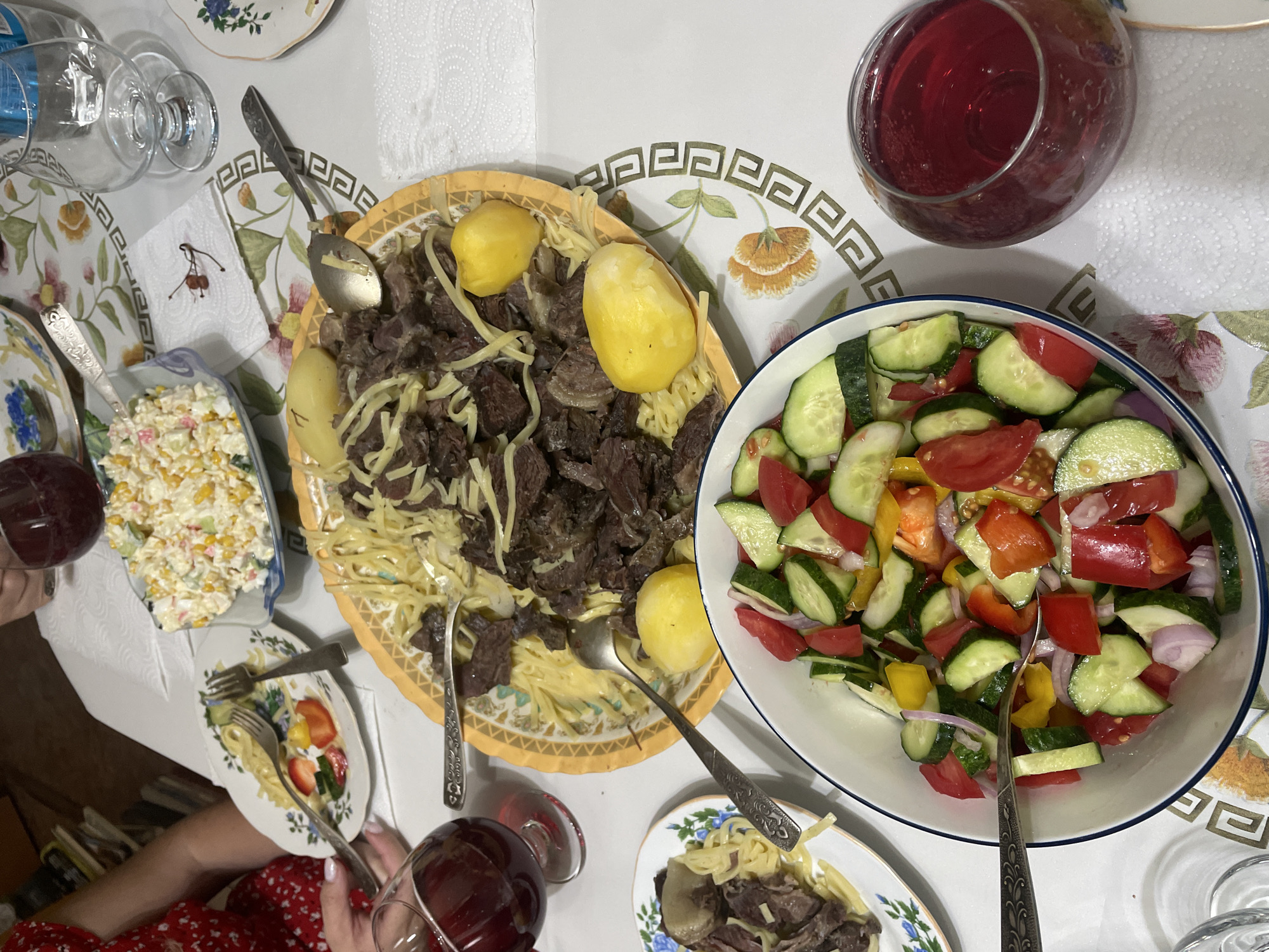 The image depicts a table laden with food, suggesting a meal is in progress or has just concluded. Several plates of food are visible, including a large platter of noodles with meat and potatoes, a bowl of chopped salad with tomatoes, cucumbers, and onions, and a smaller plate with pasta, strawberries, and cucumber. There are also glasses of red liquid, likely wine, and a bottle of water. The table is covered with a patterned tablecloth. The presence of a small, decorative napkin with a drawn violin and bow suggests a possible artistic or cultural theme associated with the gathering. The overall impression is one of a casual yet festive meal. The specific location in Almaty, Kazakhstan, cannot be determined from the image alone, as it lacks any identifying landmarks or context.