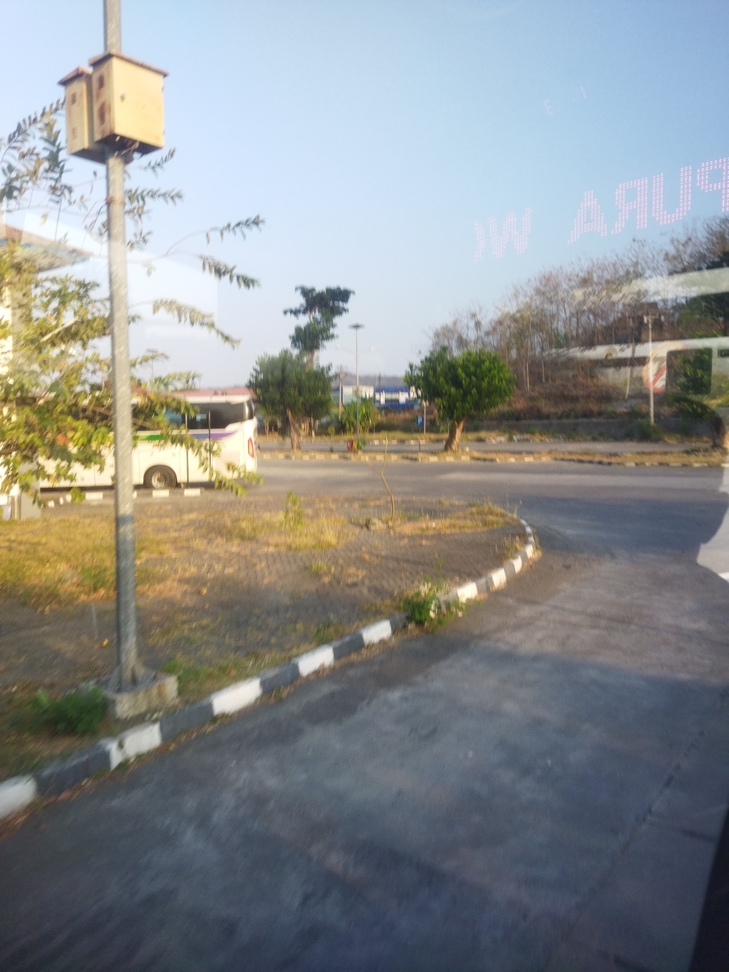 The image shows an outdoor scene with a road and some vegetation. In the foreground, there is a paved road with a curb that is painted with alternating black and white stripes. To the left of the road, there is an area of dry grass and some scattered green plants. A utility pole with a light-colored box attached to it stands prominently on the left side of the frame. Behind the utility pole, a white bus with purple and green accents is parked. Further in the background, there are trees and some buildings, including a structure with a white facade and red trim. The sky is clear and blue. Reflections of what appear to be text are visible on the sky, suggesting the photo may have been taken through a window. The overall environment appears to be an urban or semi-urban area. The location is Singodutan, Indonesia.