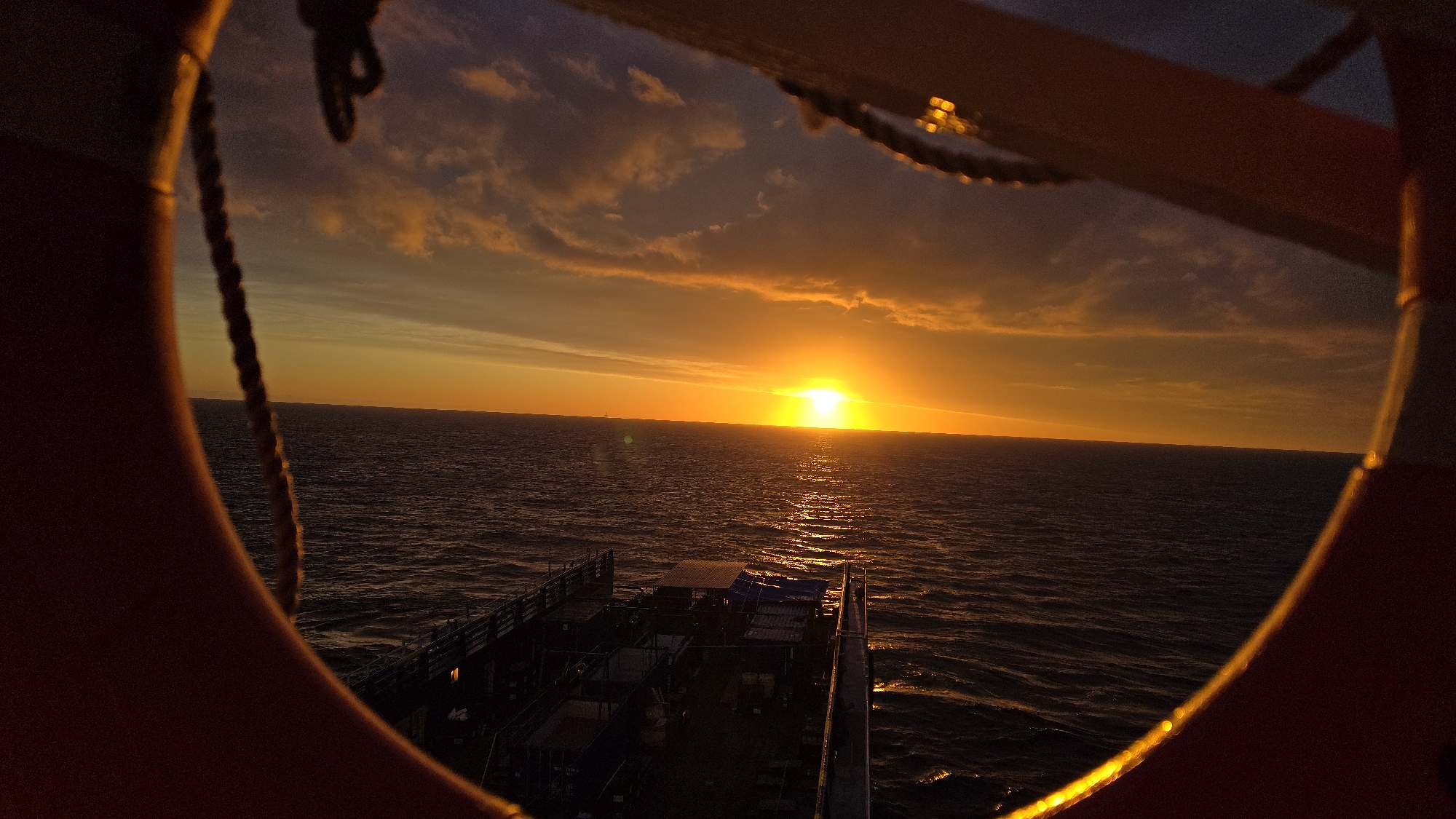 The image depicts a sunset over the ocean, as seen through a porthole of a boat. The sun is low on the horizon, casting a golden light across the water and sky. There is a boat visible in the foreground of the picture. The location is unspecified.