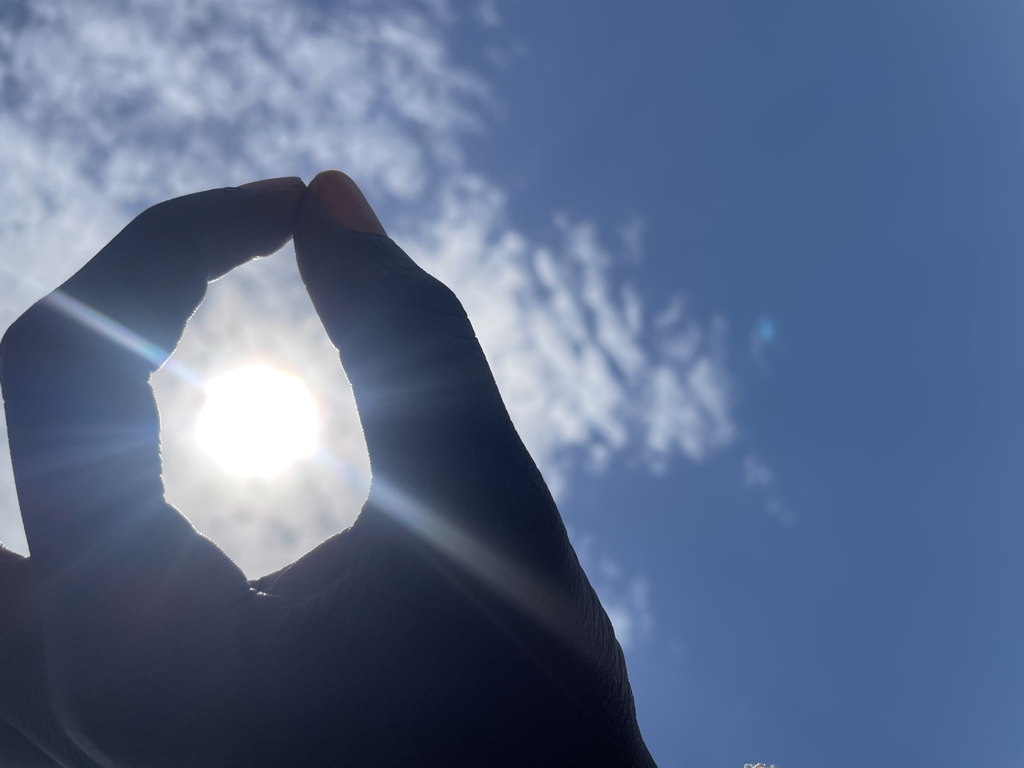 A close-up, low-angle, silhouette shot captures a dark hand forming an "O" shape against a bright, sunlit sky. The sun is positioned in the center of the "O," creating a lens flare that extends outwards. The sky is a vibrant blue with scattered white clouds. The image appears to be taken outdoors on a clear day.