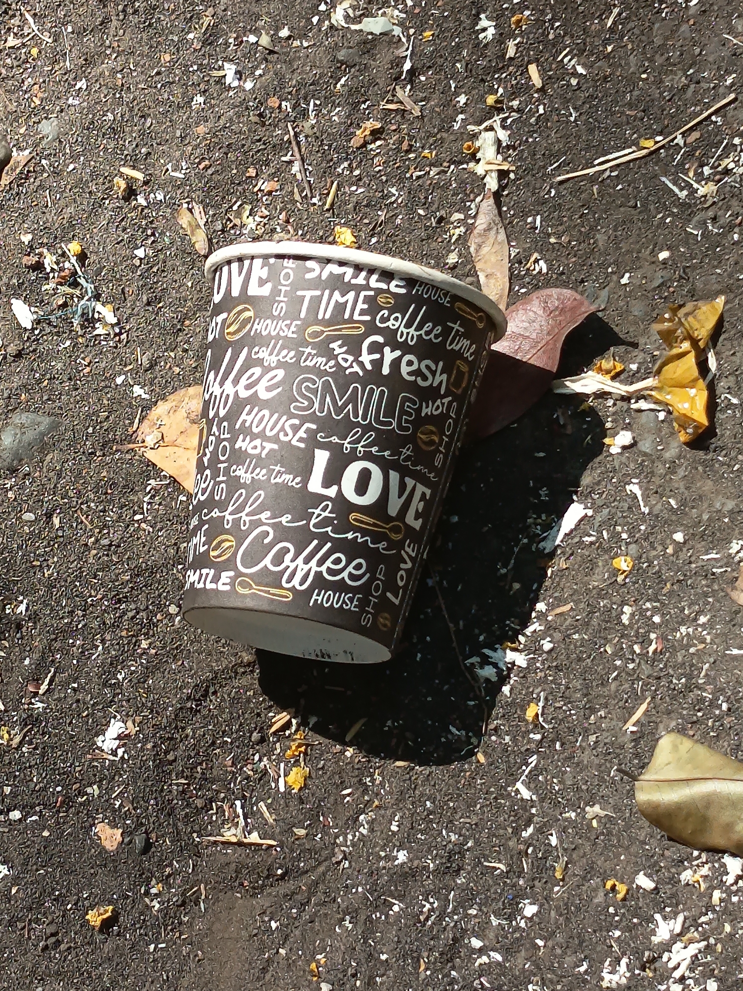 A black paper coffee cup with white text is lying on the ground in Kandangjati Kulon, Indonesia. The cup has the words "LOVE COFFEE TIME SMILE HOUSE HOT FRESH" printed on it in a repeating pattern. The ground appears to be asphalt or dirt, with scattered leaves and debris. The lighting suggests it is daytime, with a shadow cast by the cup. There are no people or animals visible in the image.