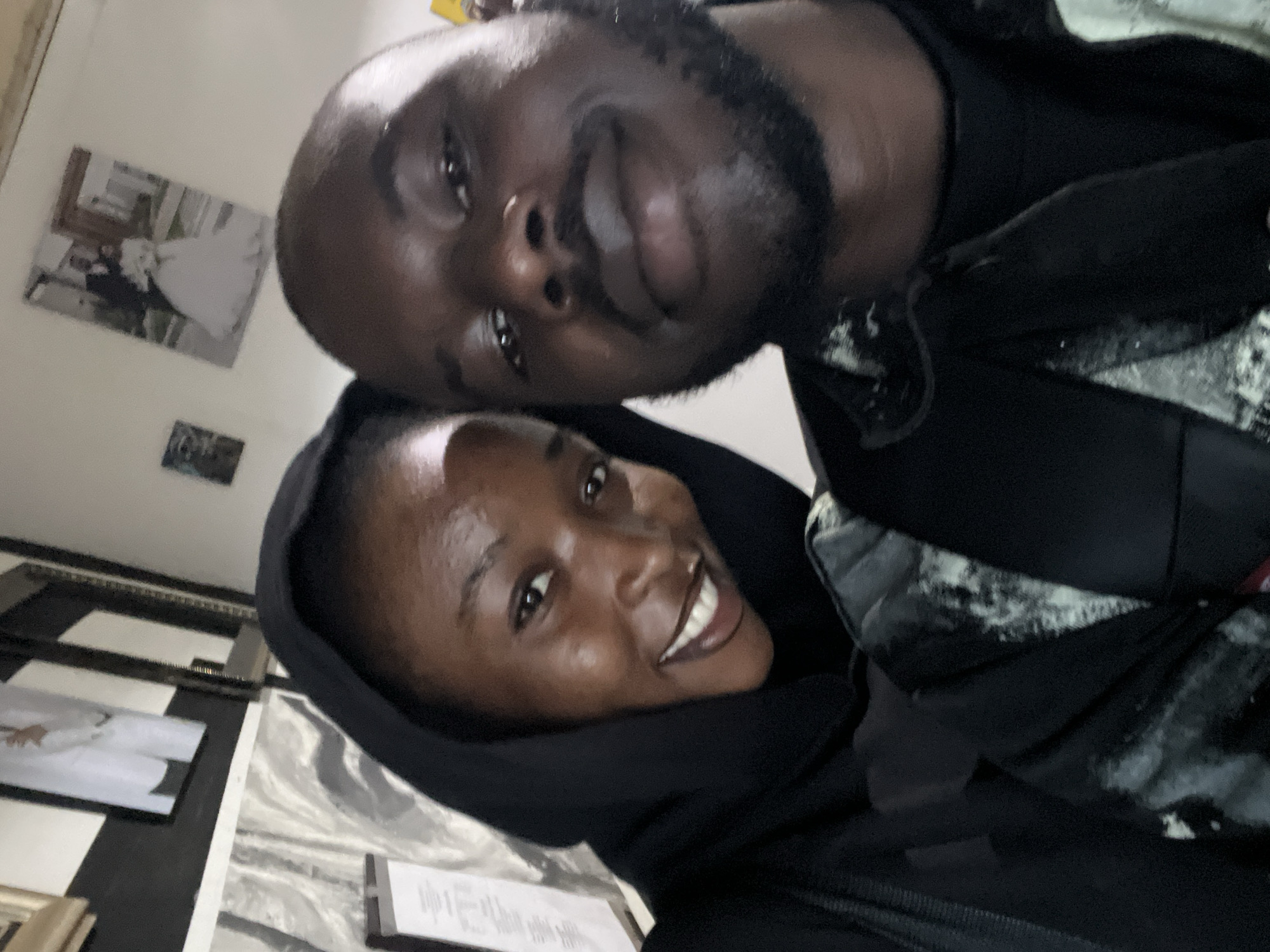 A close-up, upward-angled indoor selfie depicts two dark-skinned individuals positioned in the foreground. The individual on the left, a female, wears a black hooded garment, smiles broadly showing white teeth, and has visible moisture or sheen on her forehead and cheeks. The individual on the right, a male, has a short beard and mustache, displays a subtle smile, and wears a dark garment with an abstract grey and white pattern visible on his left shoulder. Both individuals are looking directly towards the camera.
The background consists of a light-colored wall adorned with framed pictures. To the upper left, a blurry, vertically oriented framed image shows a figure in white attire. To the upper right, another blurry, vertically oriented framed image depicts a wedding couple, with the man in a dark suit and the woman in a white wedding gown. Below and to the left of the female subject, a partial view of a clipboard or document with illegible text is visible. The lighting appears to be artificial, casting some highlights and shadows on the subjects' faces. The specific location within Jos, Nigeria, is not visually discernible from the image content.