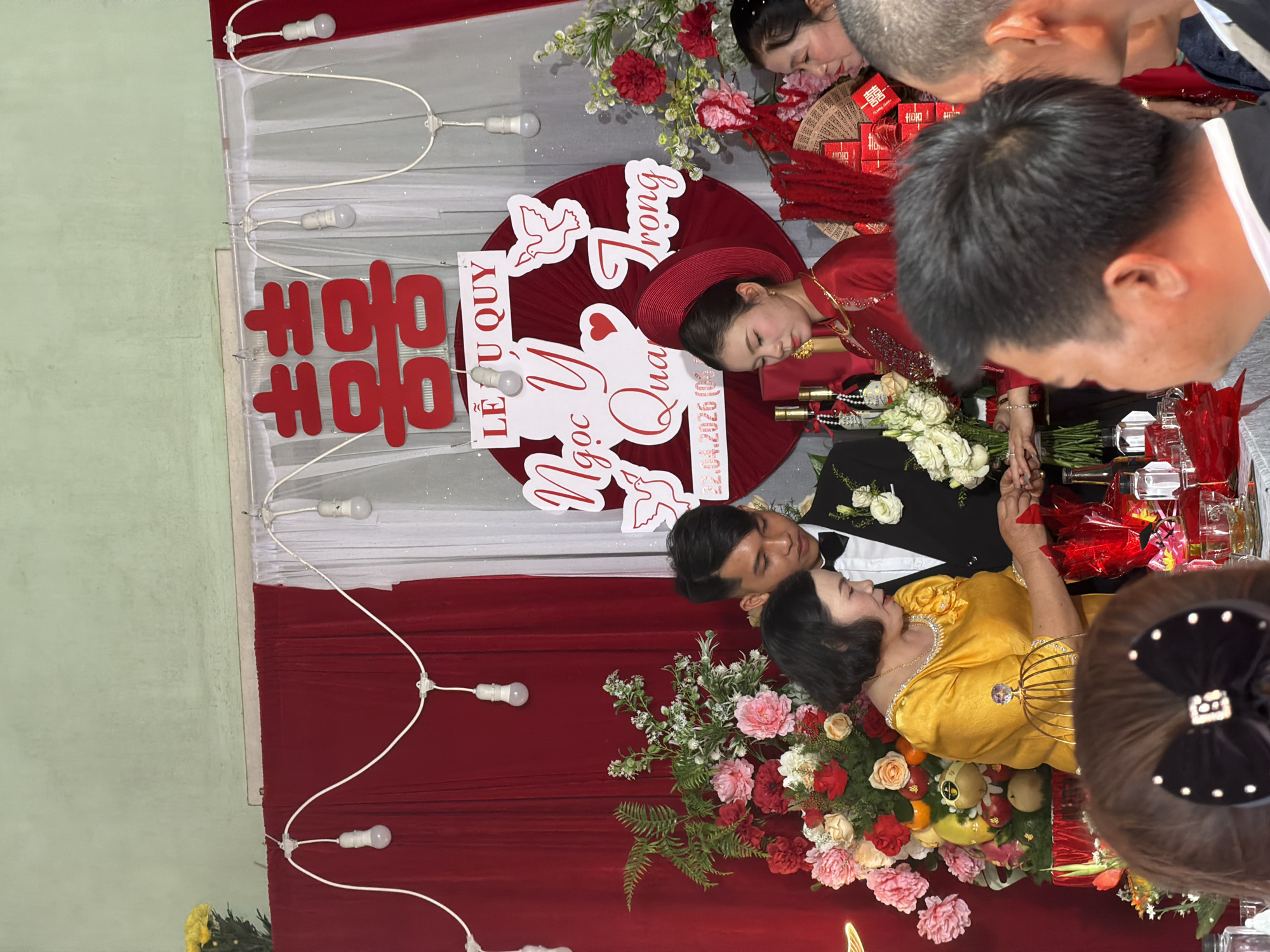A wedding scene unfolds in Tay Son Commune, Vietnam. The backdrop features red and white curtains adorned with string lights. The wedding sign reads "Le U Quy Ngoc Y Qua Trung". The bride and groom lie on the table, dressed in traditional Vietnamese attire. The table is lavishly decorated with flowers, fruit, and bottles. Family members are gathered around, observing the ceremony.