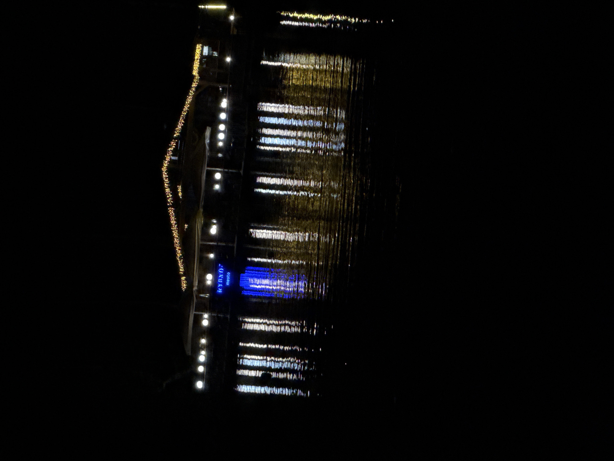 The image depicts a nighttime scene at Zolotoi Plyazh, Russia, featuring a structure illuminated by decorative lights, likely a restaurant or entertainment venue, situated on the water. The structure is adorned with strings of yellow lights along its roofline and is further illuminated by white lights. A prominent blue sign with the text "RYBNOE mesto" is visible on the facade of the structure. The lights from the structure are reflected on the dark, rippling water surface, creating a shimmering effect. The overall environment is dark, suggesting the presence of a body of water at night. No individuals or specific activities are discernible.