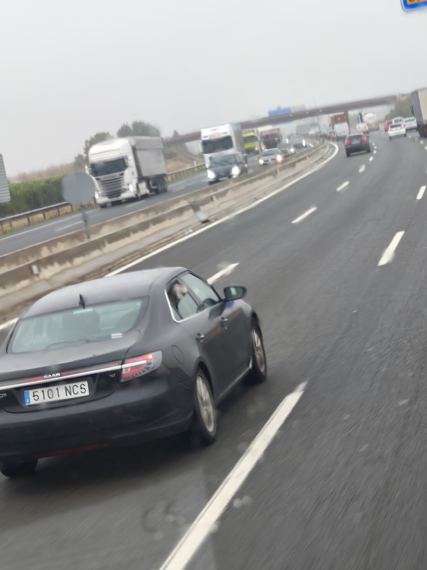 A dark grey Saab sedan is captured from a rear-left perspective, driving on a wet, multi-lane highway under an overcast sky. A person is visible in the driver's seat. The vehicle displays a Spanish license plate "5101 NCS" and "SAAB" and "TiD" badging on its trunk.
The highway is busy with moderate to heavy traffic, suggesting slow-moving conditions or congestion. In the lane to the left of the Saab, a white Scania truck is clearly visible, followed by other trucks and various cars and vans extending into the distance. An overhead bridge spans the highway in the background, further along the road.
The wet road surface indicates recent rain or ongoing precipitation, contributing to the overall gloomy and muted lighting of the scene. Guard rails separate the highway from green vegetation on the side. The image portrays a typical highway setting, consistent with the provided location context of Quart de Poblet, Spain, and the visible Spanish license plate.