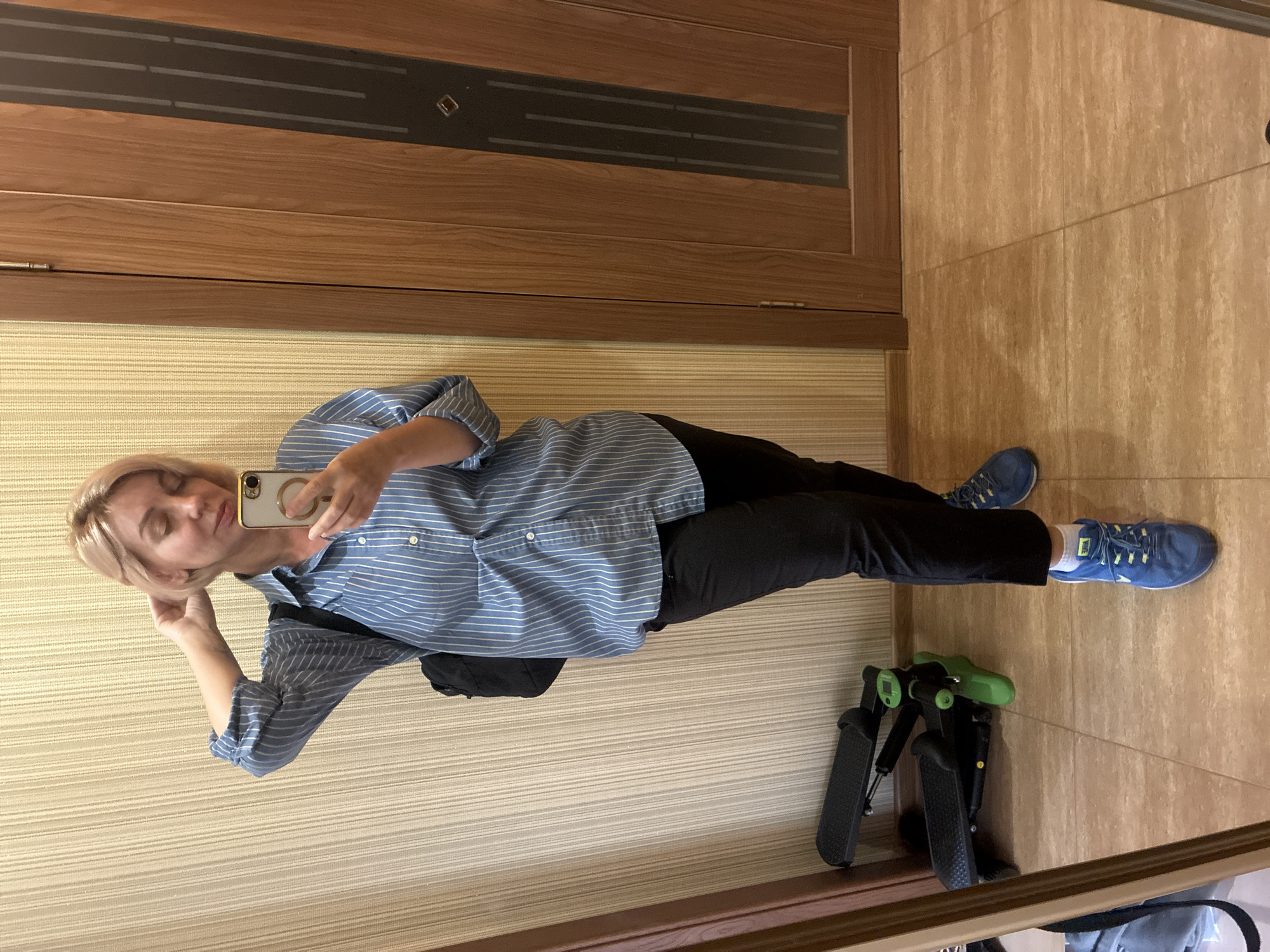 A Caucasian woman with short blonde hair is standing in an indoor space, taking a mirror selfie. She is wearing a blue and white striped button-down shirt, black pants, and blue athletic shoes with white socks. She has a black backpack slung over her left shoulder and is holding a smartphone in her right hand, which is raised to touch her hair. Her eyes are closed and her head is tilted.
The environment appears to be an apartment or house. The background features a wall with vertical textured wallpaper in a neutral tone, and a wooden door with vertical paneling on the right side of the frame. In the lower left corner, a black and green stair stepper exercise machine is visible. The floor is tiled in a light brown, tan, and beige pattern. The lighting suggests an indoor environment, possibly with artificial light. There is no indication of the city of Sochi, Russia, in the image.