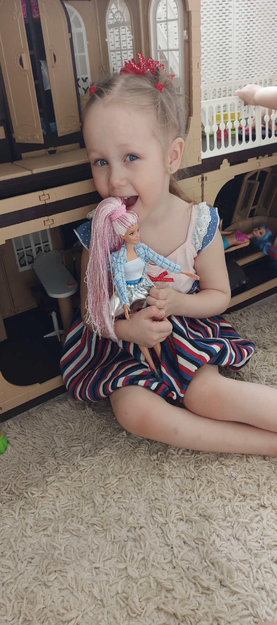 A young girl with fair skin, blue eyes, and blond hair pulled into small pigtails secured with red hair ties and a large red bow, sits cross-legged on a beige, high-pile carpet. She is smiling broadly, holding a Barbie doll with long pink hair. The girl is wearing a striped dress with alternating red, white, and blue stripes, a light pink tank top with ruffled lace straps.
Behind the girl is a large, multi-story dollhouse with arched windows and decorative detailing. Inside the dollhouse, other dolls are visible. Another child's hand reaches into the doll house, which makes us believe the picture was taken in her home in Chaikovsky, Russia.