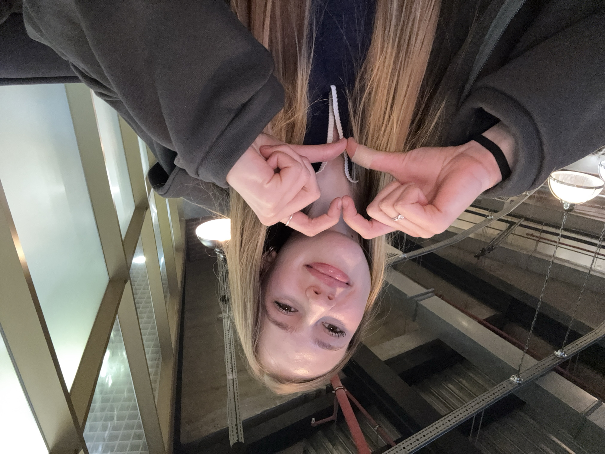 A young, light-skinned female with long, straight, blonde hair, parted centrally, smiles directly at the camera. Her tongue is slightly extended between her lips. She wears dark eye makeup. Her hands are positioned in front of her chest, forming a heart shape with her fingers. A small, light-colored bandage is visible on the knuckle of her left index finger. She wears a silver-colored ring on her left ring finger and a simple silver-colored band on her right ring finger. A black band or bracelet is visible on her left wrist. She is dressed in a dark gray or charcoal-colored hooded garment, with a darker garment featuring white drawstrings visible underneath.
The background consists of an indoor architectural space. To the left, an exposed ceiling features metal ducts, structural beams, and reddish-brown pipes, indicative of an industrial or modern design aesthetic. Several pendant light fixtures with white, dome-shaped shades hang from the ceiling structure. To the right, a wall composed of large, light-colored framed panels, some appearing frosted or translucent, allows diffused light into the space. A geometric grid pattern is discernible through the upper sections of these panels. There are no identifiable landmarks, signs, or unique architectural features within the image to corroborate a specific location in Sinyavino, Russia.