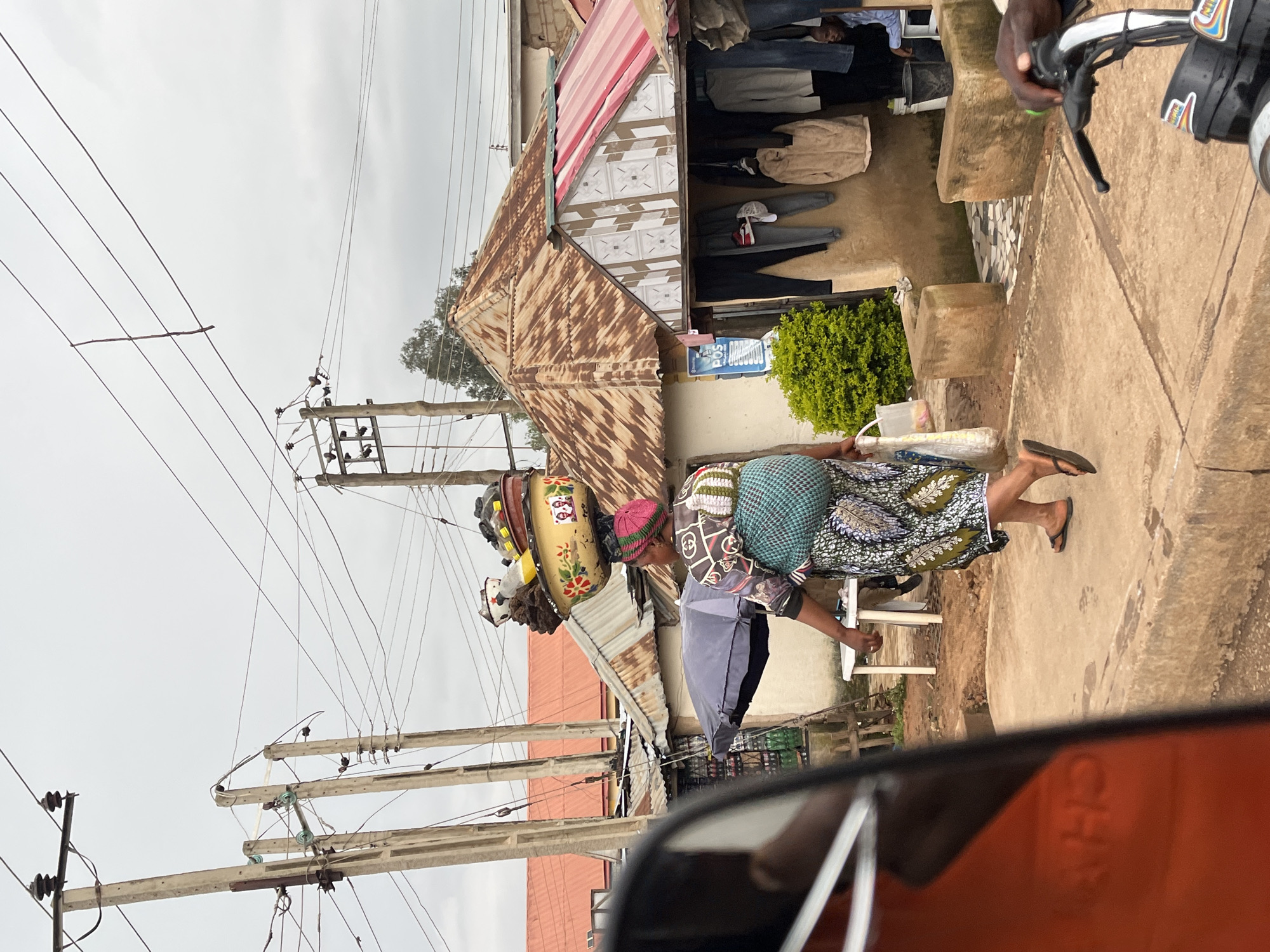 A woman is seen from behind, carrying a large basket of items on her head and a baby on her back, walking along a dirt and concrete path. She is wearing a patterned dress and a green cloth wrapped around the baby. The background shows a street scene in Bukuru, Nigeria, with several buildings and power lines overhead. One building has a rusty, corrugated metal roof and an awning with red and white stripes. Clothes are hanging on display outside another structure. In the foreground, a portion of a vehicle's orange side mirror is visible.